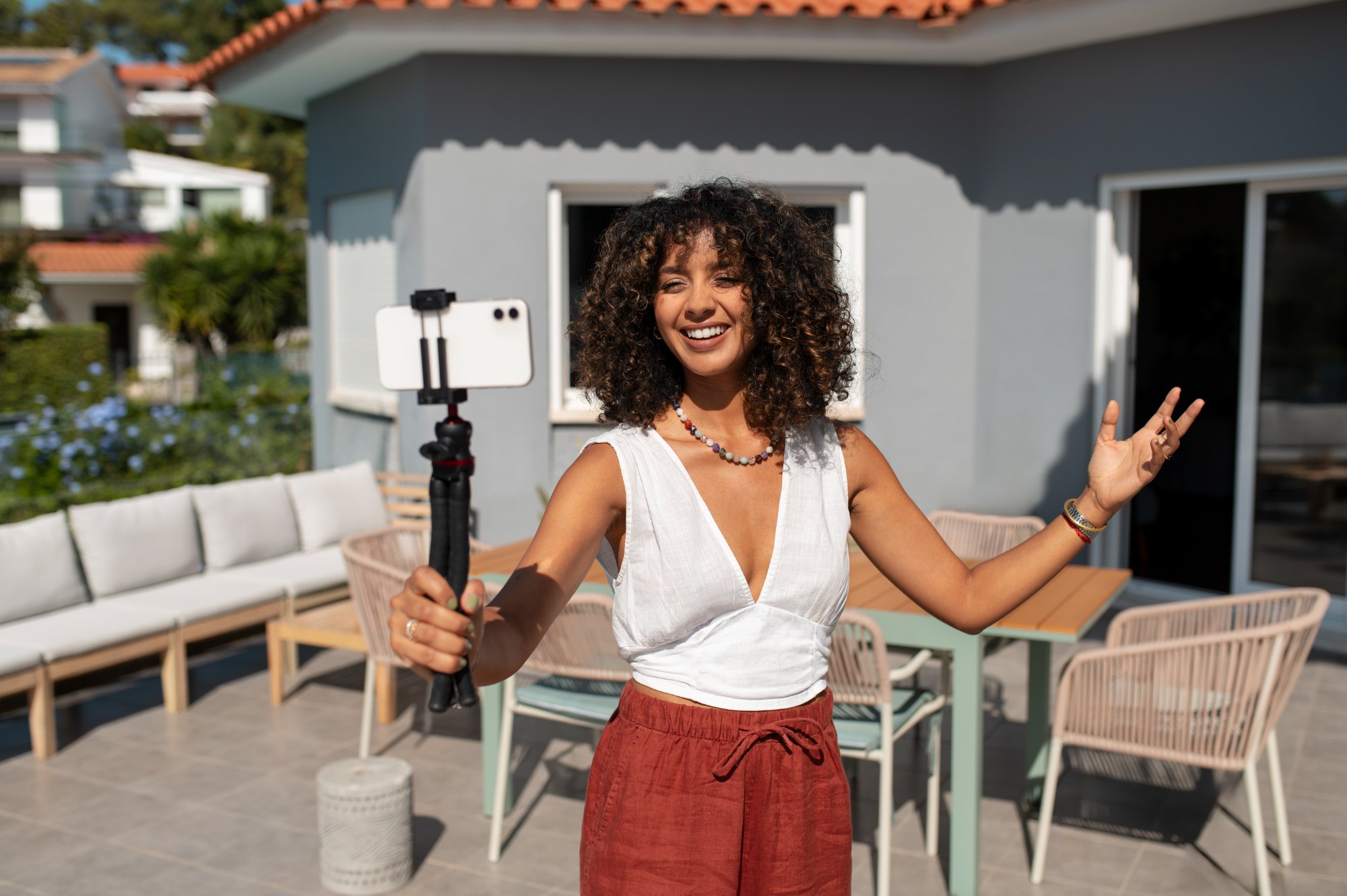 Mulher de cabelo cacheado grava vídeo com celular em tripé no terraço de uma casa