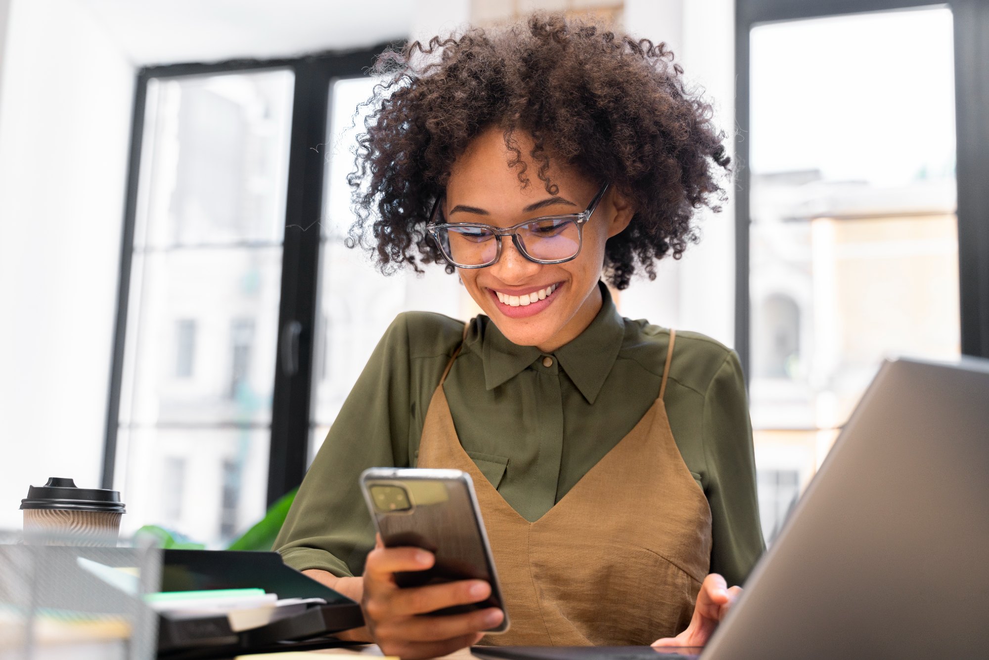 Mulher de cabelo cacheado e óculos sorri olhando o celular em escritório, com notebook e copo de café na mesa