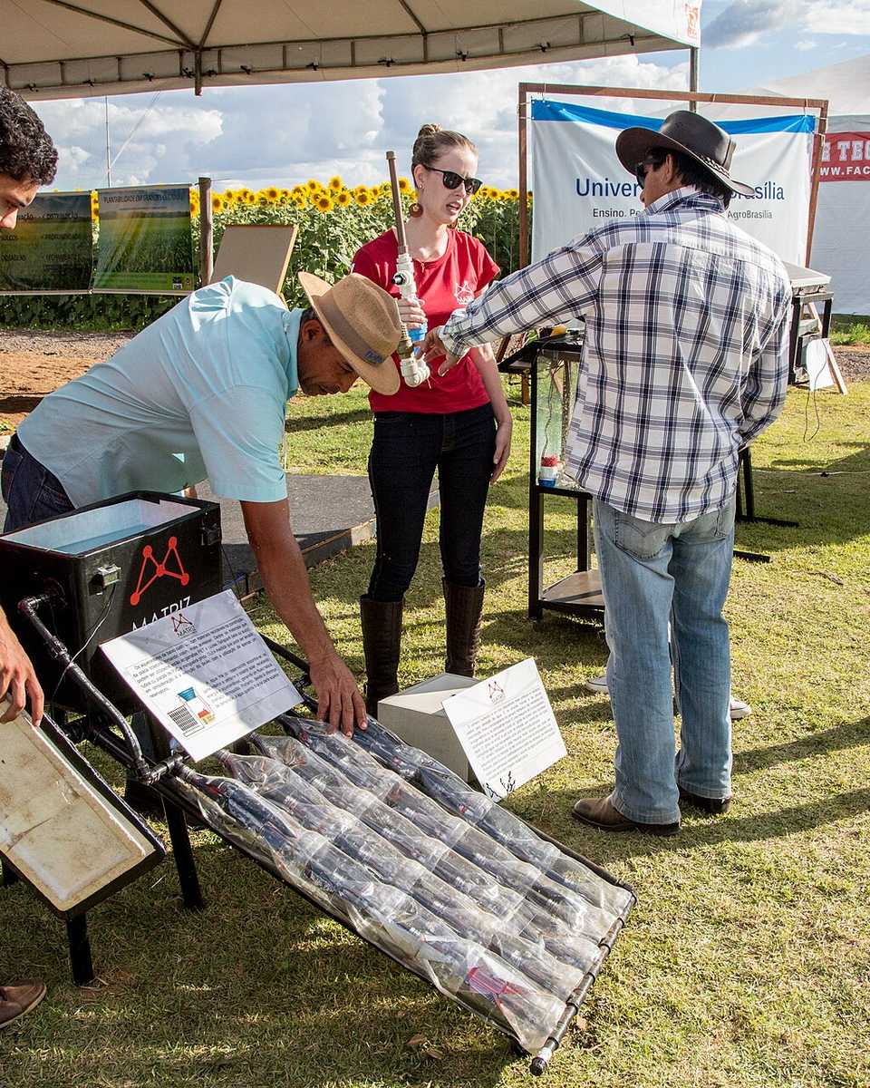 Produtores rurais de chapéu e estudantes em volta de um equipamento com painel solar montado no gramado de um estande da AgroBrasília, no PAD-DF, com tendas da feira e lavoura de girassol ao fundo.