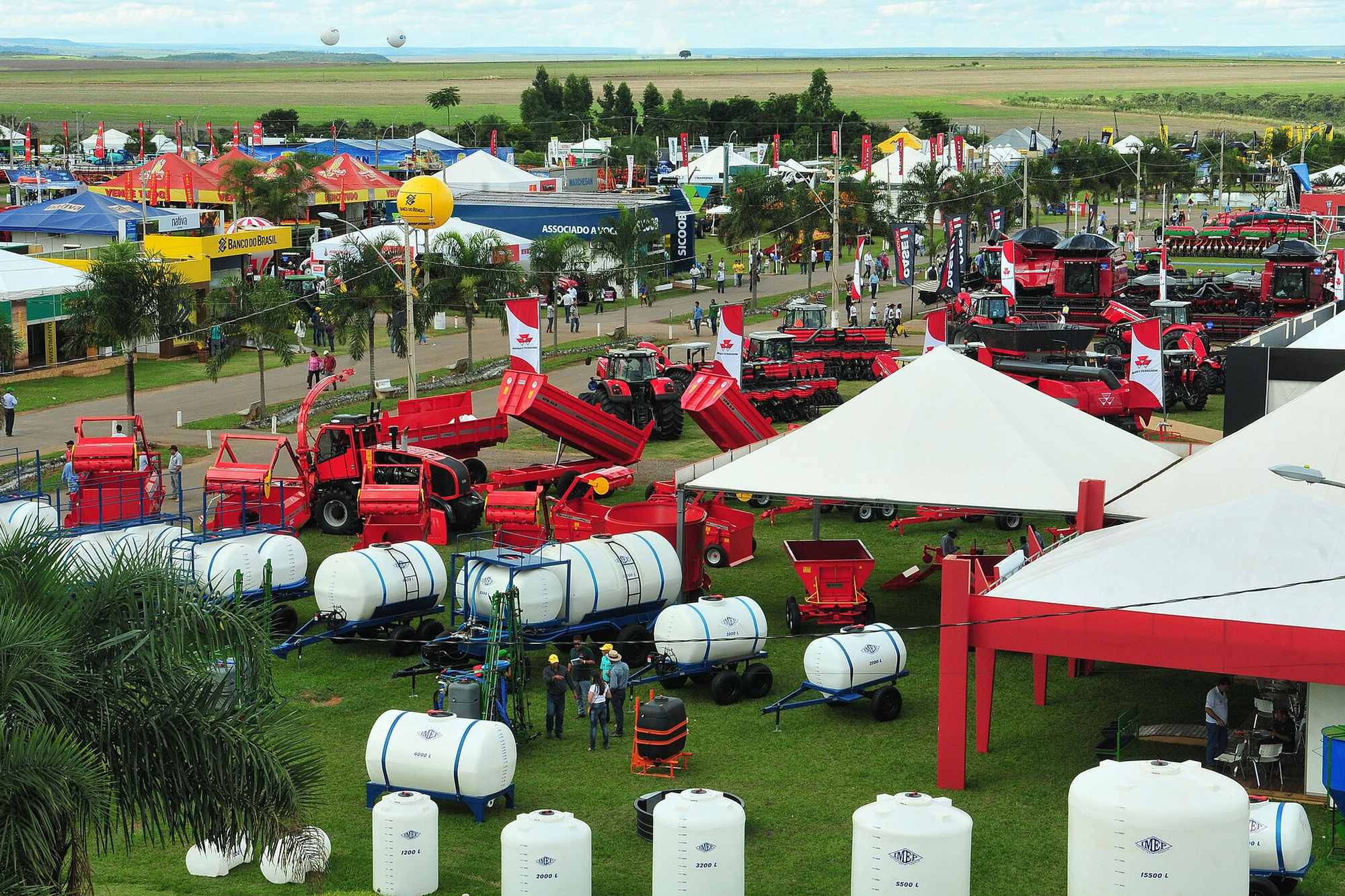 Vista do alto do parque da AgroBrasília, no PAD-DF, com fileiras de tratores e implementos vermelhos, tanques brancos, tendas de expositores e visitantes circulando entre os estandes, com a lavoura do cerrado no horizonte.