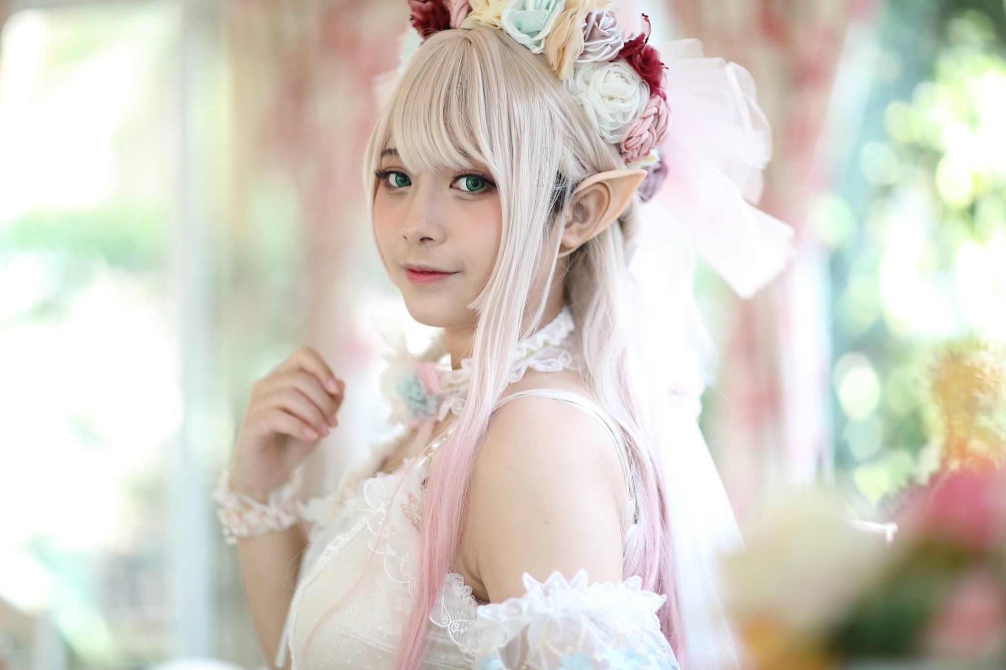 Cosplayer de peruca platinada com pontas rosa, orelhas de elfo e coroa de flores, de vestido branco de renda, posando num jardim florido.