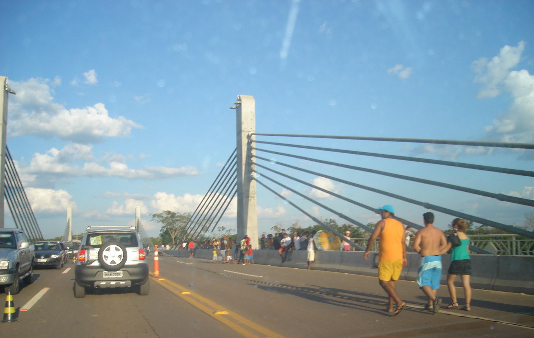Pessoas caminhando sobre a Terceira Ponte de Rio Branco ao entardecer, com carros parados na pista ao lado e a estrutura estaiada da ponte ao fundo.