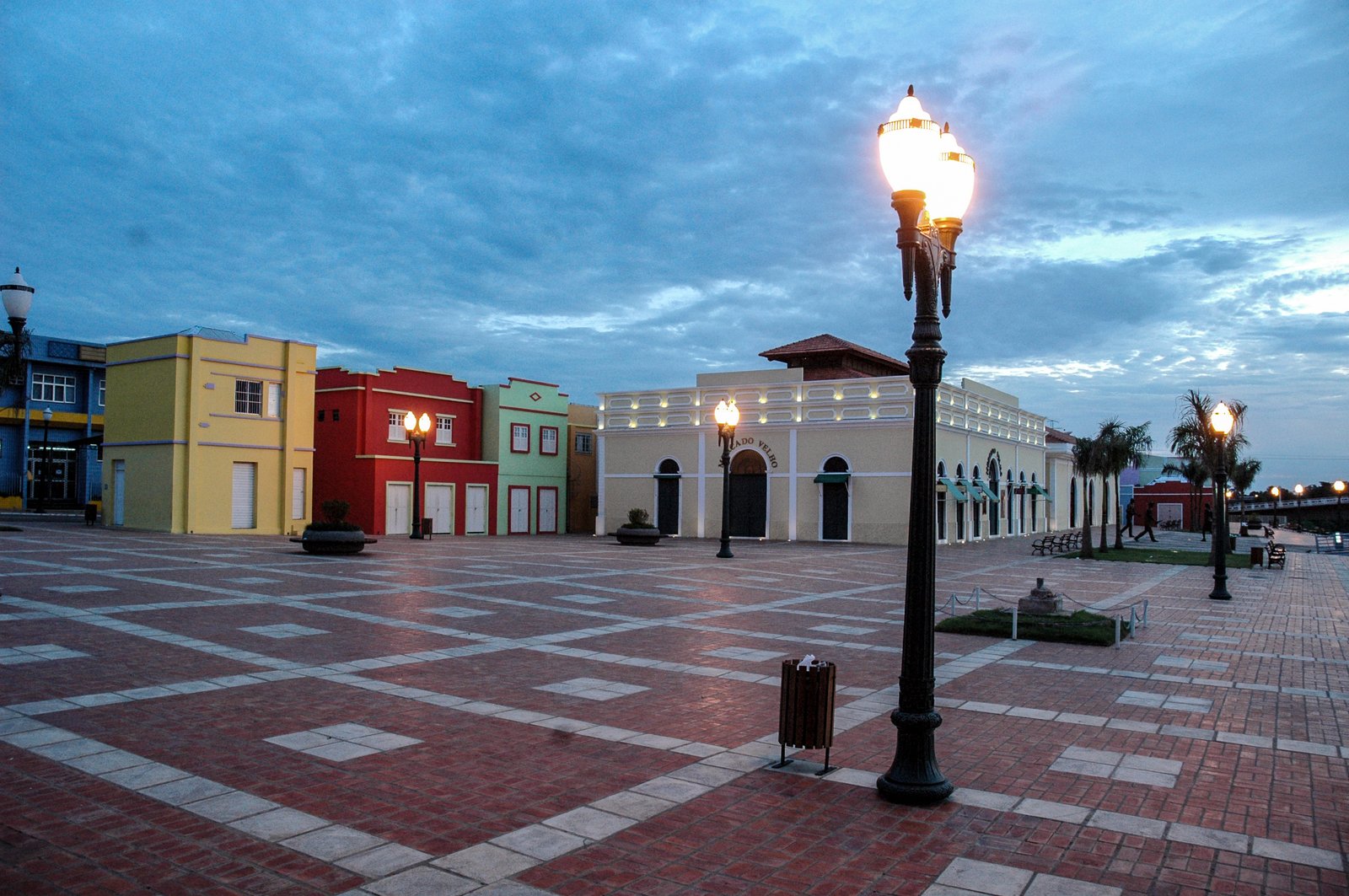 Calçadão da Gameleira ao amanhecer em Rio Branco, com os prédios coloridos do Mercado Velho iluminados por postes de luz sob um céu azul de nuvens ao entardecer.