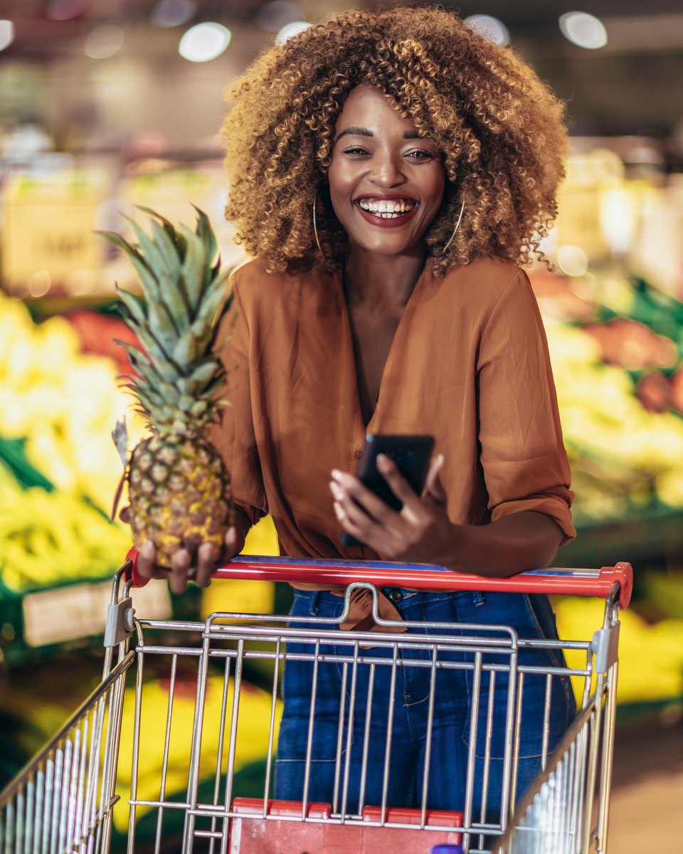 Mulher de cabelo cacheado sorrindo, apoiada num carrinho de supermercado, com um abacaxi numa mão e o celular na outra, diante da banca de frutas.