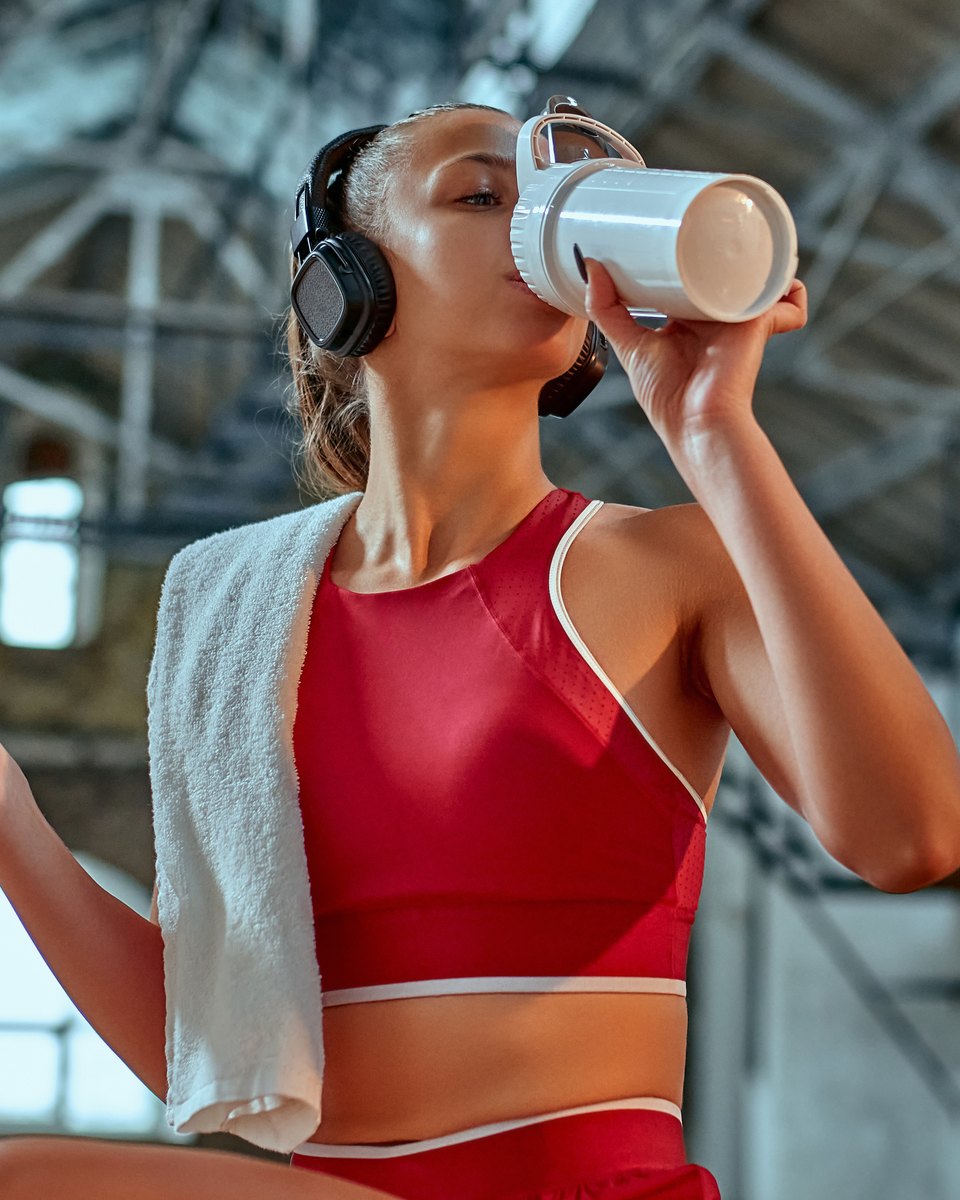 Mulher de top vermelho e fone de ouvido bebendo shake numa garrafa branca enquanto segura o celular, sentada numa academia com teto de estrutura metálica.