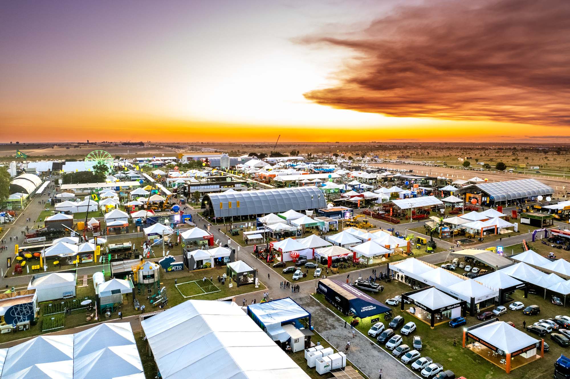 Vista aérea do parque da Bahia Farm Show, em Luís Eduardo Magalhães, ao pôr do sol, com tendas brancas, pavilhões, máquinas agrícolas, carros expostos e uma roda-gigante, e o cerrado plano do Oeste baiano até o horizonte.