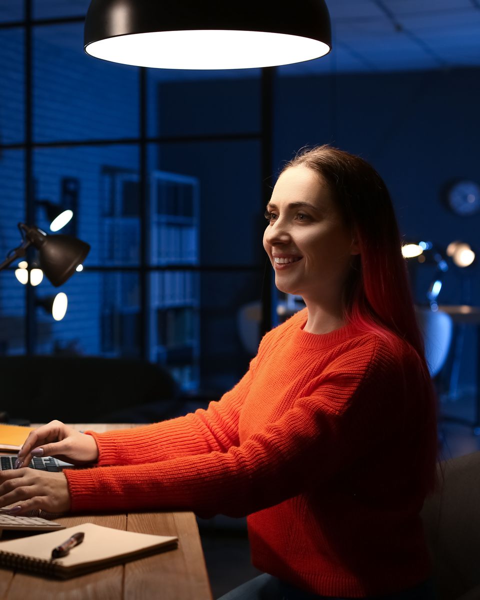 Mulher de suéter laranja sorrindo enquanto digita num notebook, numa mesa de escritório à noite, com um monitor mostrando linhas de código ao lado.