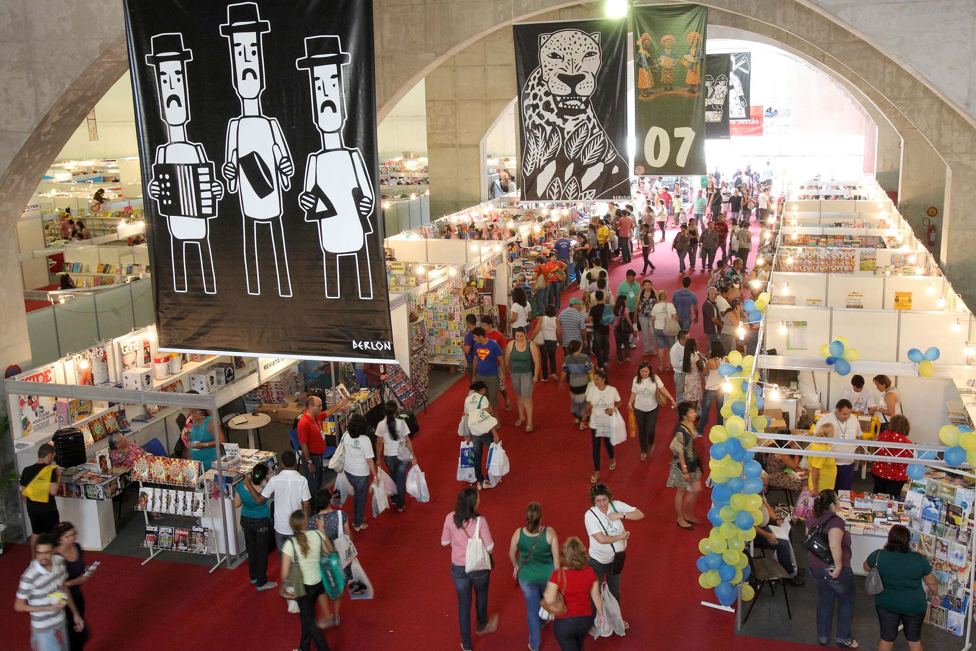 Corredor lotado da Bienal do Livro de Pernambuco visto do alto, com tapete vermelho, estandes de editoras dos dois lados, balões e grandes banners em preto e branco com figuras em estilo de xilogravura pendurados no teto.