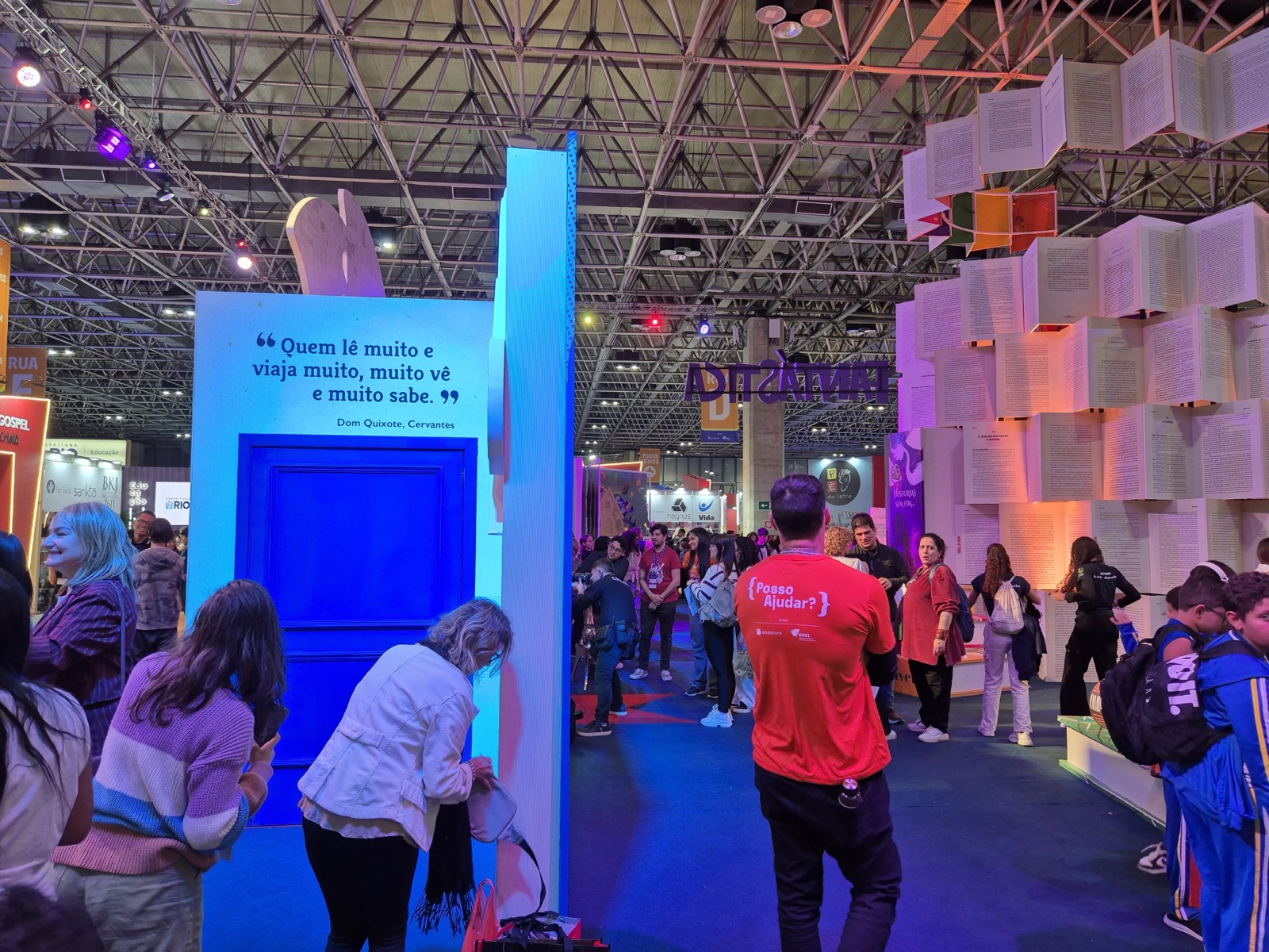 Corredor da Bienal do Livro do Rio de Janeiro em 2025 com luz azul, visitantes fotografando uma grande porta azul com a frase de Dom Quixote "Quem lê muito e viaja muito, muito vê e muito sabe", uma parede de livros abertos à direita e um voluntário de camiseta vermelha de costas.
