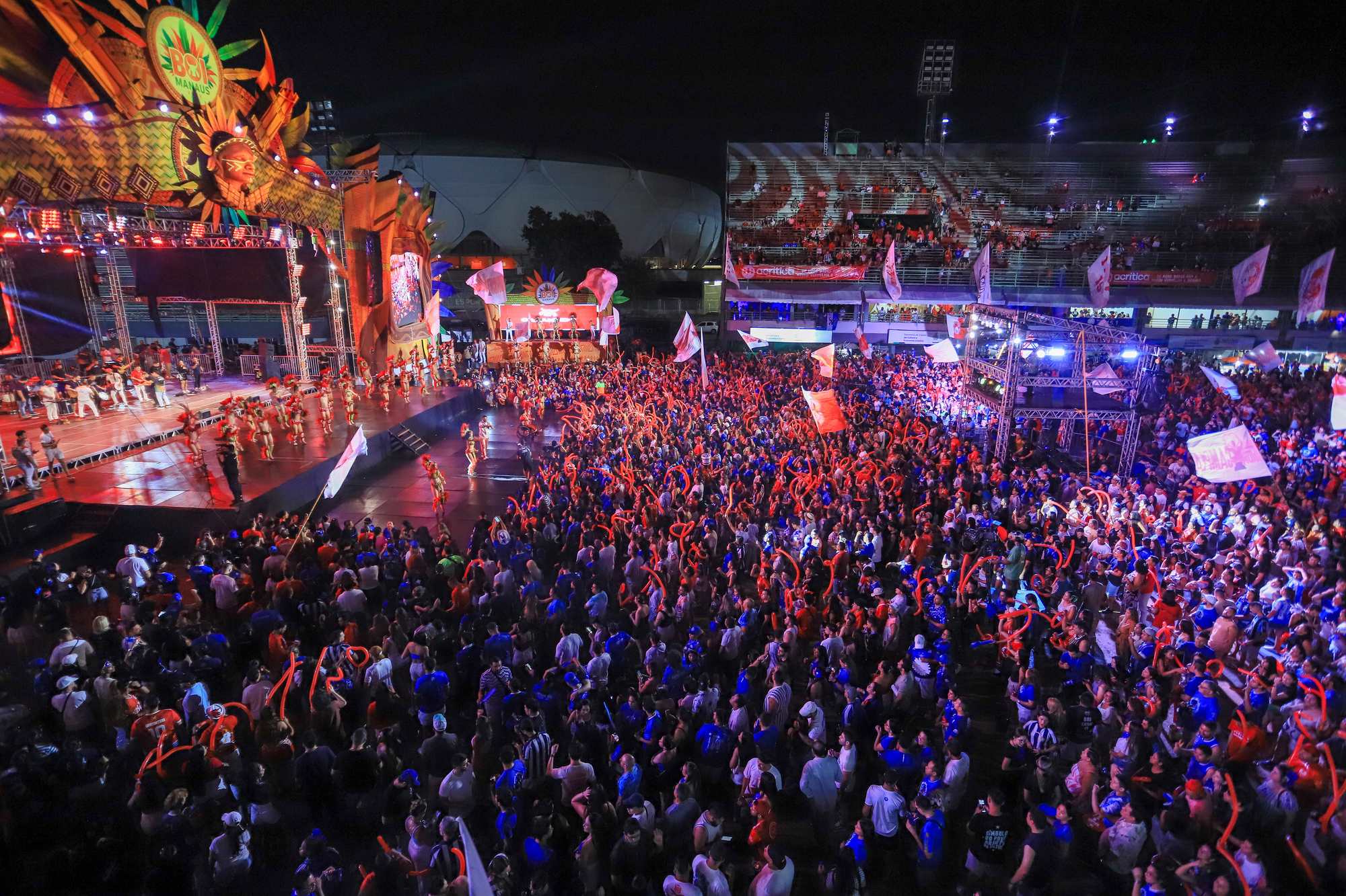 Público lotando o Sambódromo de Manaus à noite no Boi Manaus, com balões vermelhos e azuis misturados na multidão, o palco iluminado à esquerda e a Arena da Amazônia ao fundo.