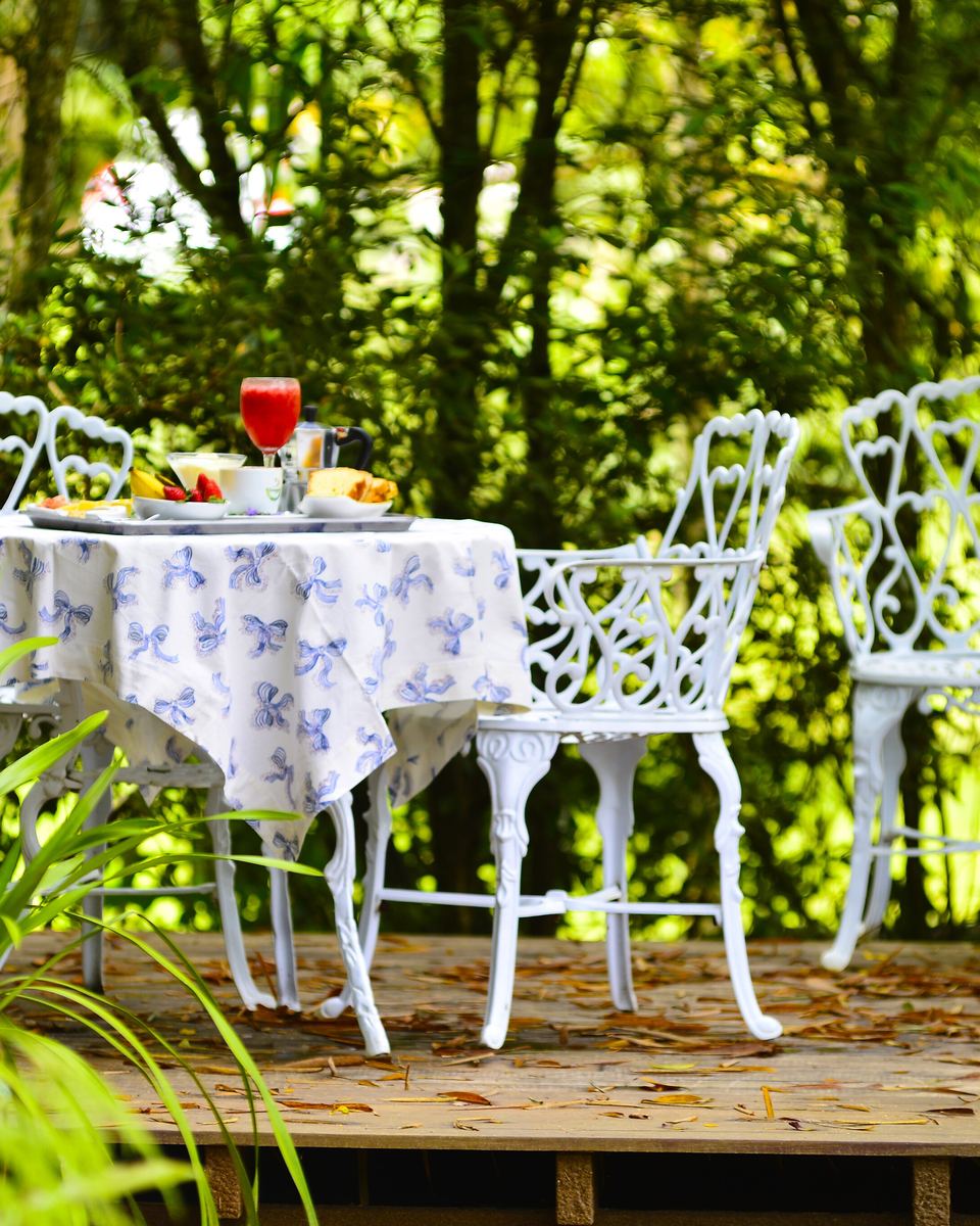 Mesa de café da manhã posta ao ar livre numa propriedade rural de Domingos Martins, com toalha estampada, cadeiras de ferro branco, frutas, pães e suco, cercada de mata.