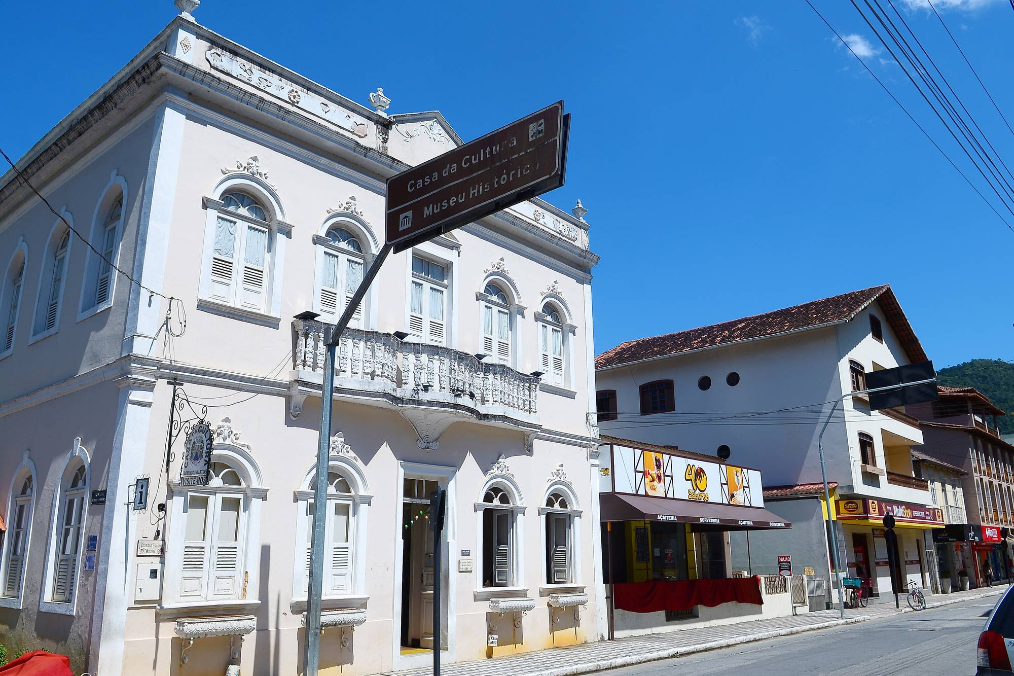 Rua do centro de Domingos Martins em dia de sol, com o casarão branco da Casa da Cultura e Museu Histórico na esquina, comércio ao lado e morros verdes ao fundo.