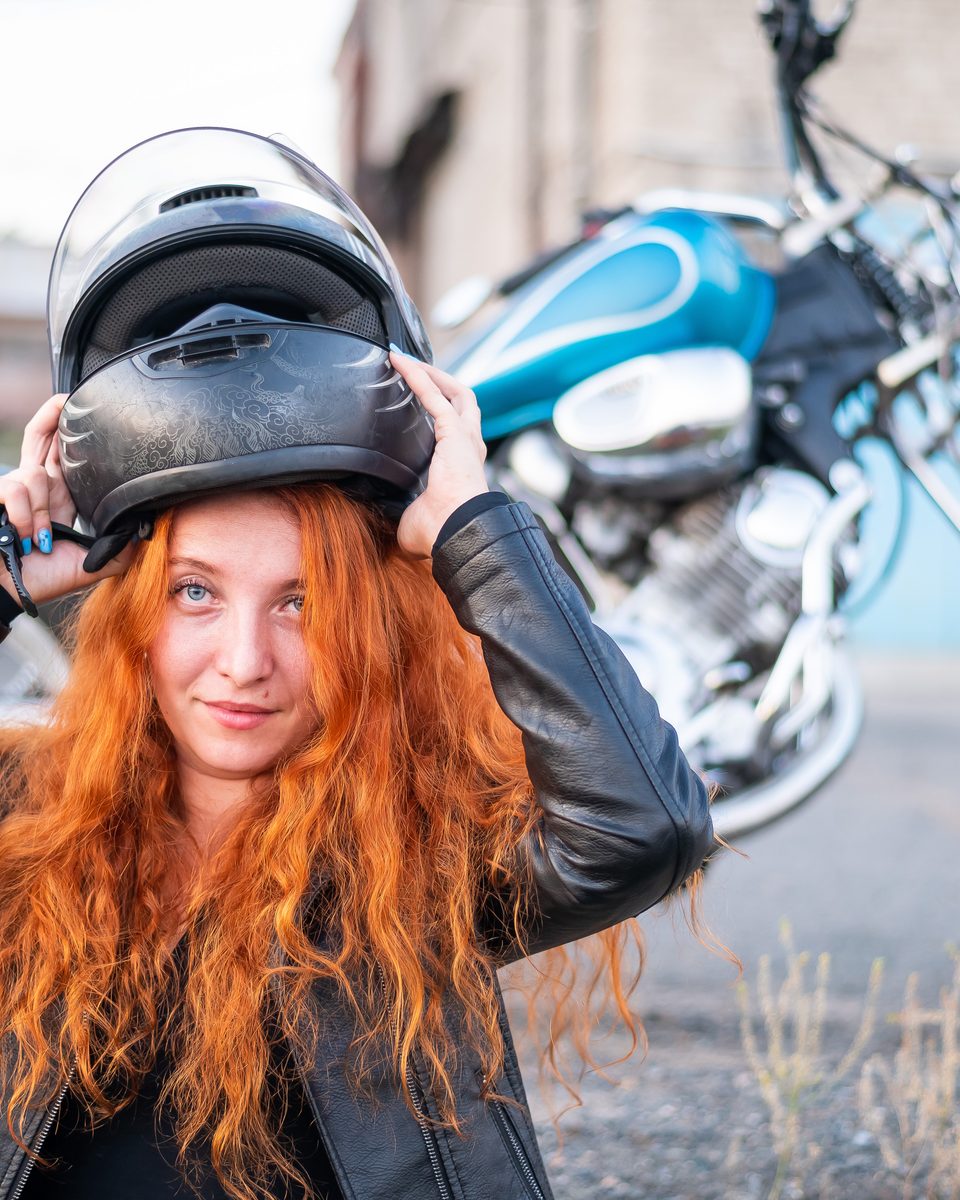 Motociclista ruiva de jaqueta de couro preta colocando o capacete, com uma moto custom azul de guidão alto estacionada atrás dela.