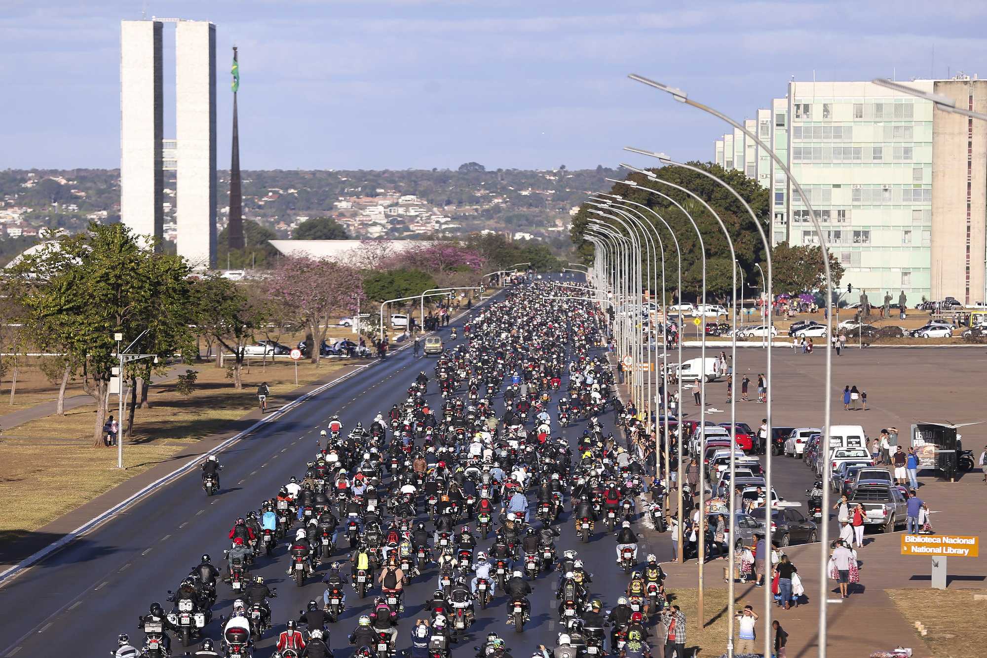 Milhares de motociclistas de capacete ocupando as pistas do Eixo Monumental, em Brasília, no passeio de encerramento do Capital Moto Week, com as torres do Congresso Nacional ao fundo, ipês floridos e público assistindo na calçada.