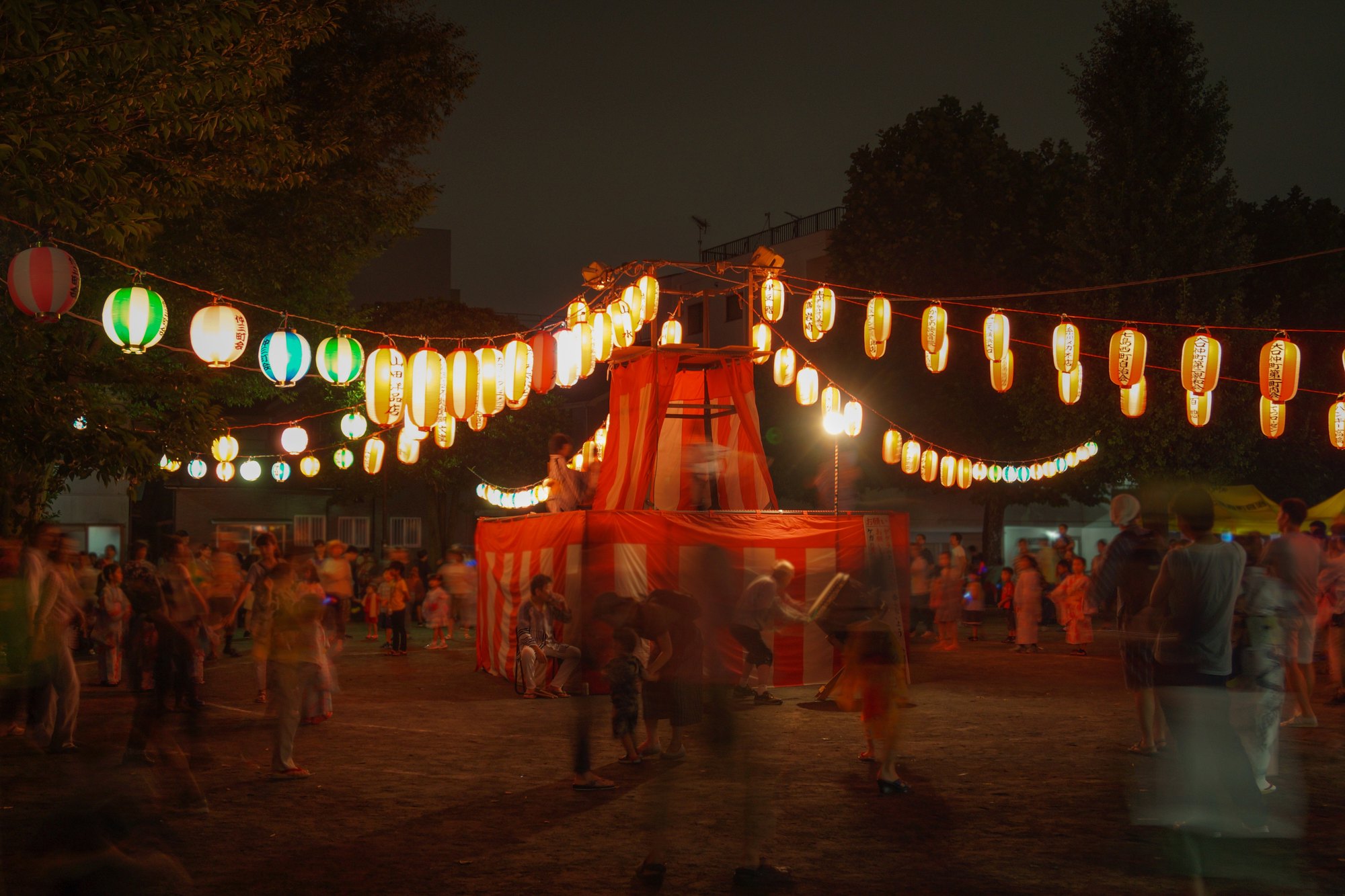 Festival de verão japonês à noite, com fileiras de lanternas de papel acesas em volta de uma torre listrada de vermelho e branco e pessoas dançando em roda.