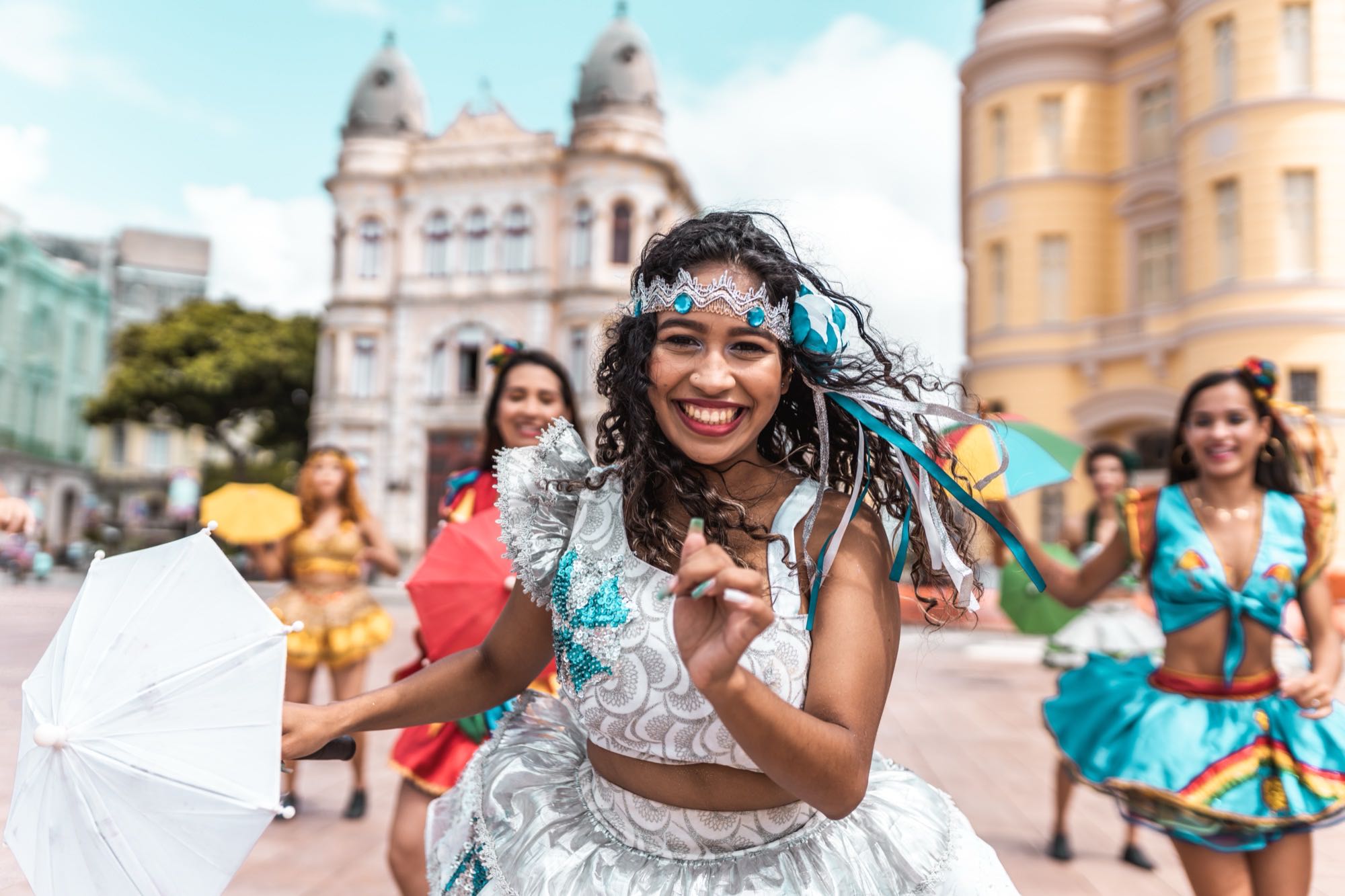 Dançarina de frevo sorrindo e dançando com uma sombrinha branca numa rua de carnaval, com outras dançarinas de saia colorida ao fundo.