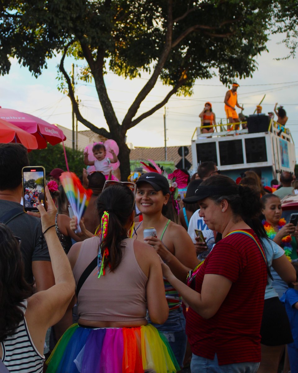 Foliã de braço erguido filmando com o celular um bloco de bairro no Graça, em Belo Horizonte, entre mulheres de saia de tule colorida, leque de arco-íris e o carro de som ao fundo.