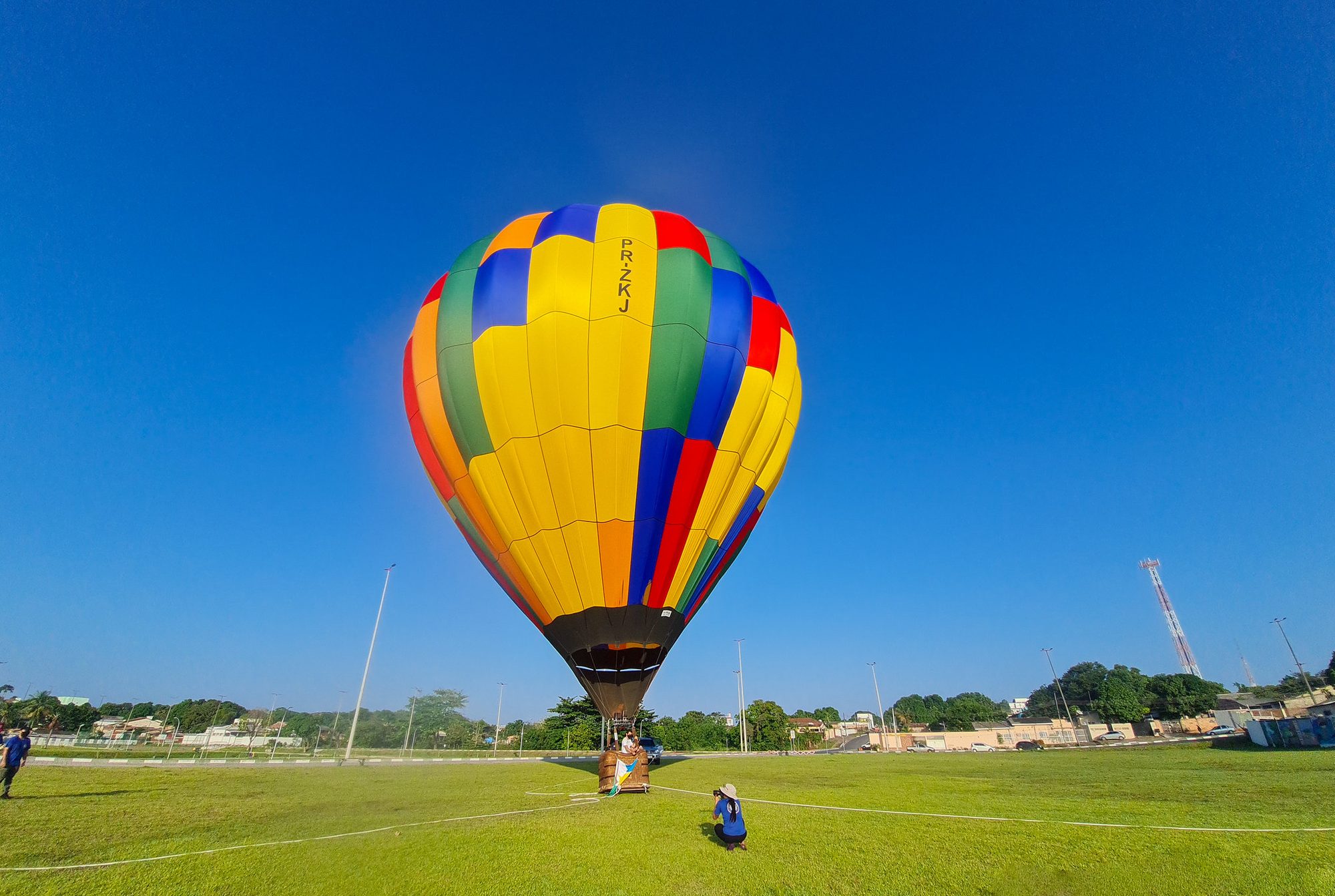 Balão de ar quente colorido, em amarelo, verde, azul e vermelho, preparado para decolar num gramado do Parque Rio Branco em Boa Vista, com uma pessoa agachada fotografando ao lado, sob céu azul sem nuvens.