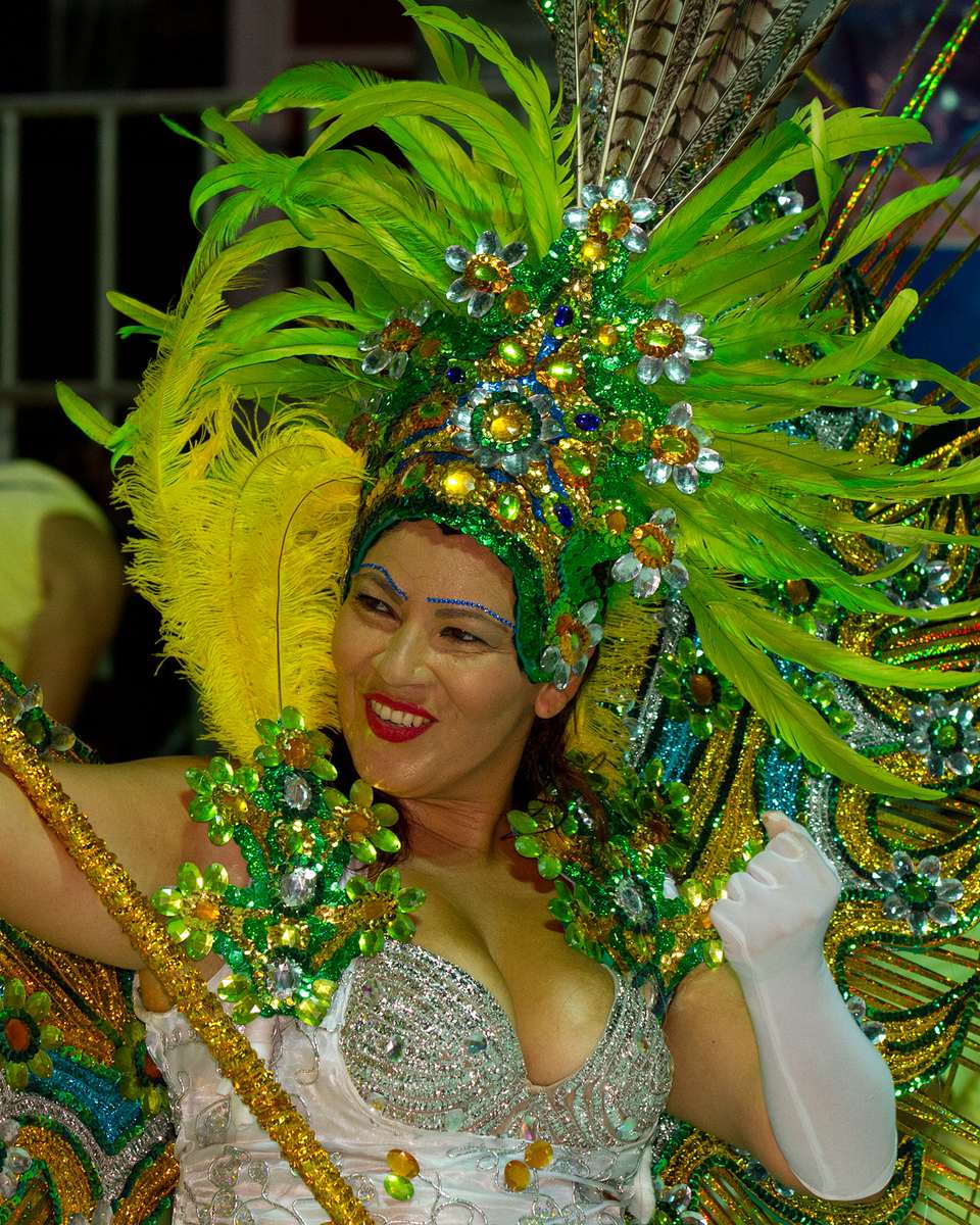 Passista sorrindo com fantasia de plumas verdes e luvas brancas durante o desfile do Carnaval de Corumbá, com o público espremido atrás dela.