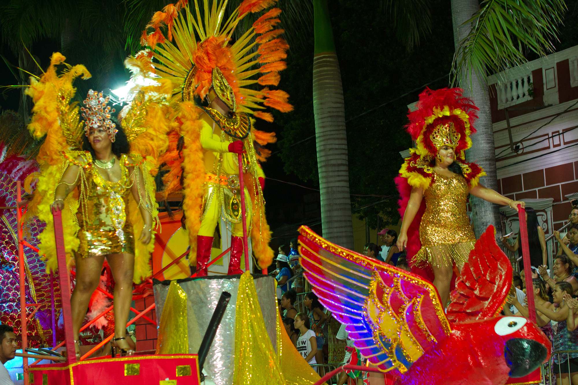 Carro alegórico vermelho e dourado com passistas de plumas amarelas e vermelhas desfilando à noite em Corumbá, com o público atrás das grades e o casario antigo ao fundo.