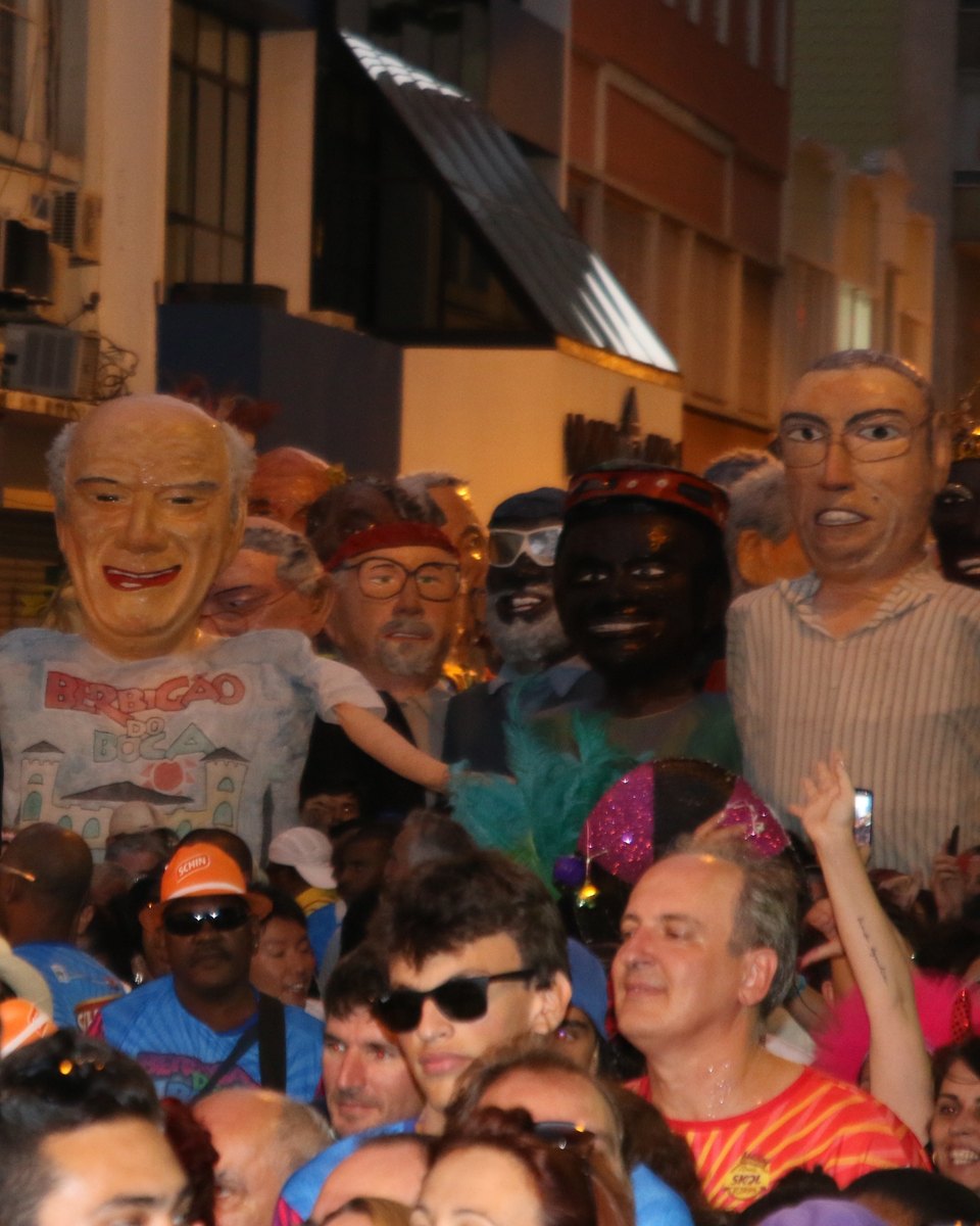 Bonecos gigantes do Berbigão do Boca, um deles com camiseta do bloco, sobre a multidão de foliões numa rua do Centro de Florianópolis, à noite, com prédios iluminados ao fundo.