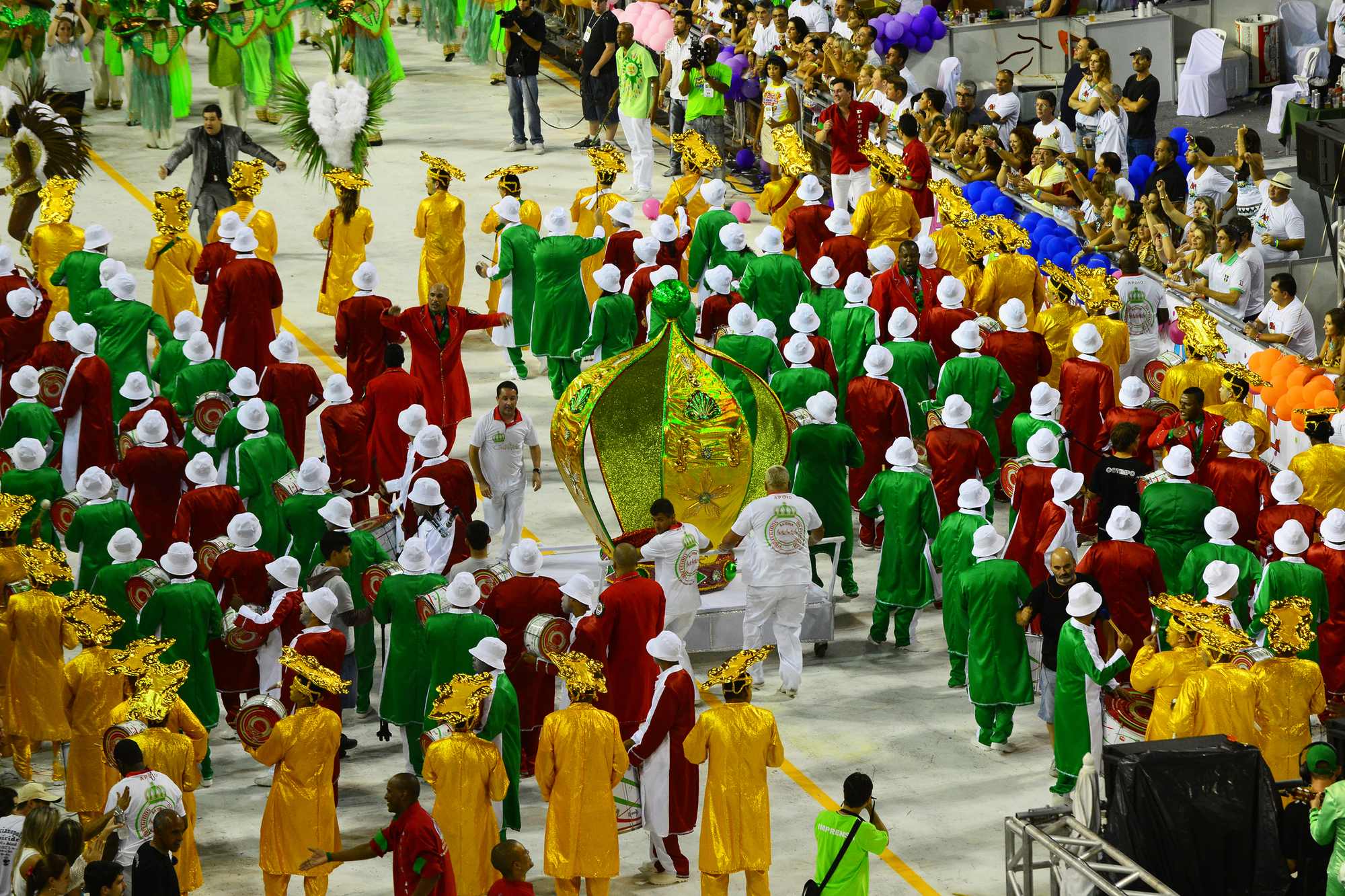 Bateria de escola de samba de roupas verdes, vermelhas e douradas, com chapéus brancos, desfilando na Passarela Nego Quirido, em Florianópolis, com a arquibancada lotada à direita.
