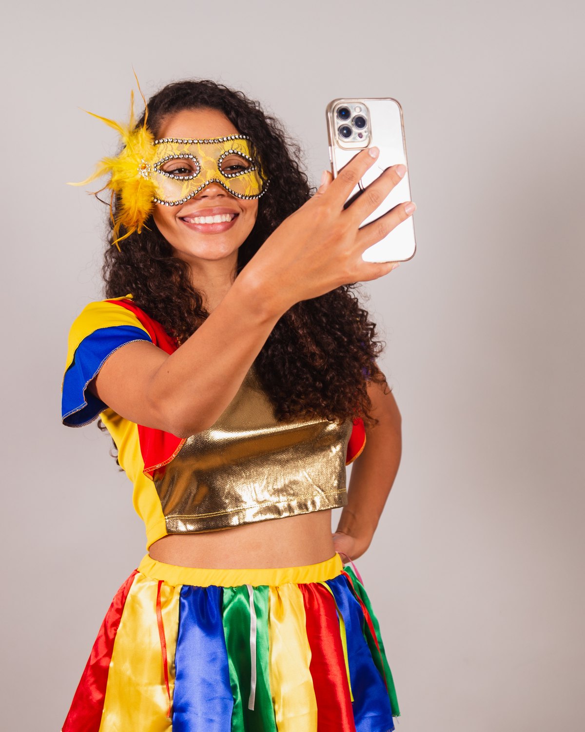 Mulher de cabelo cacheado com máscara dourada de plumas, top dourado e saia listrada de cores vivas, sorrindo enquanto grava um vídeo de si mesma com o celular.