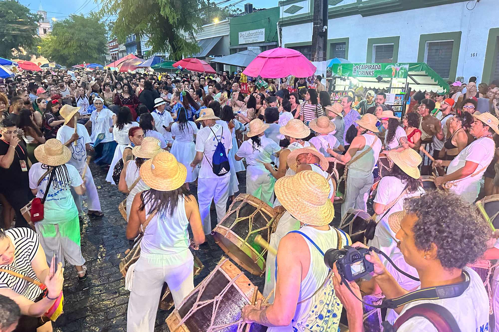 Bloco descendo uma rua do Centro Histórico de João Pessoa com percussionistas de chapéu de palha e roupa branca tocando zabumba, guarda-sóis abertos e casario colonial dos dois lados.