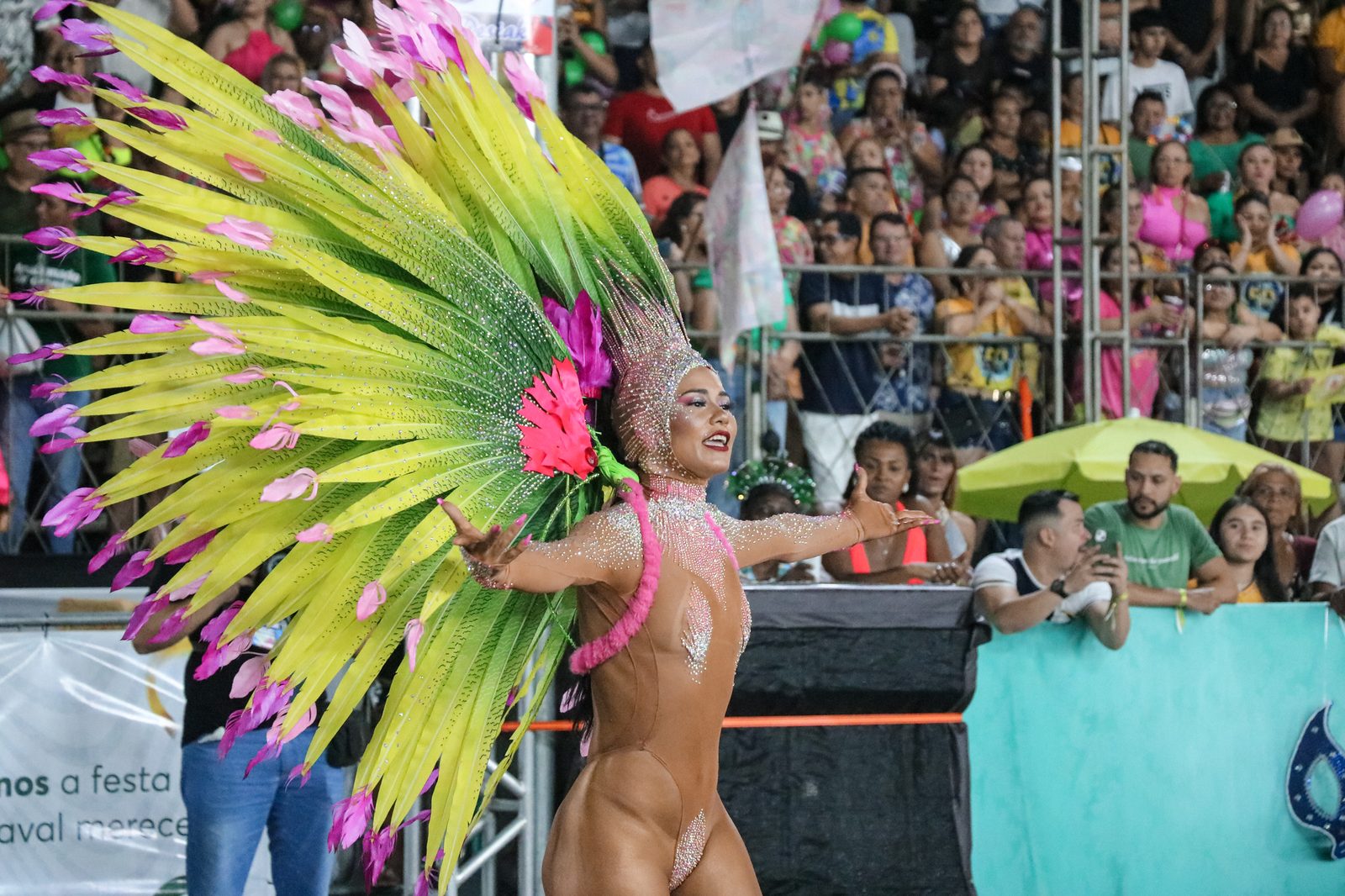 Passista dançando na avenida do Sambódromo de Macapá, com grande adereço de penas amarelas e rosa na cabeça, braços abertos, e arquibancada lotada de público ao fundo.