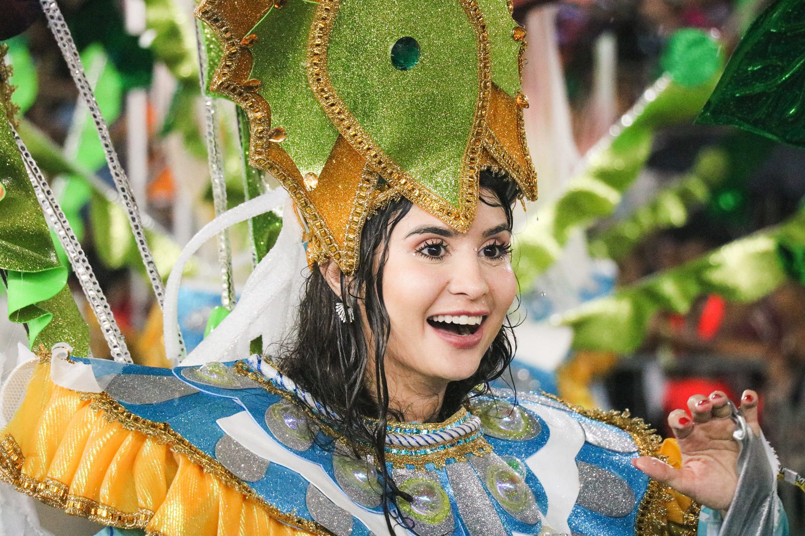 Passista sorrindo de perto, com tiara dourada e verde em forma de flor, cabelo molhado e fantasia azul e dourada com babado amarelo no ombro, em desfile no Sambódromo de Macapá.