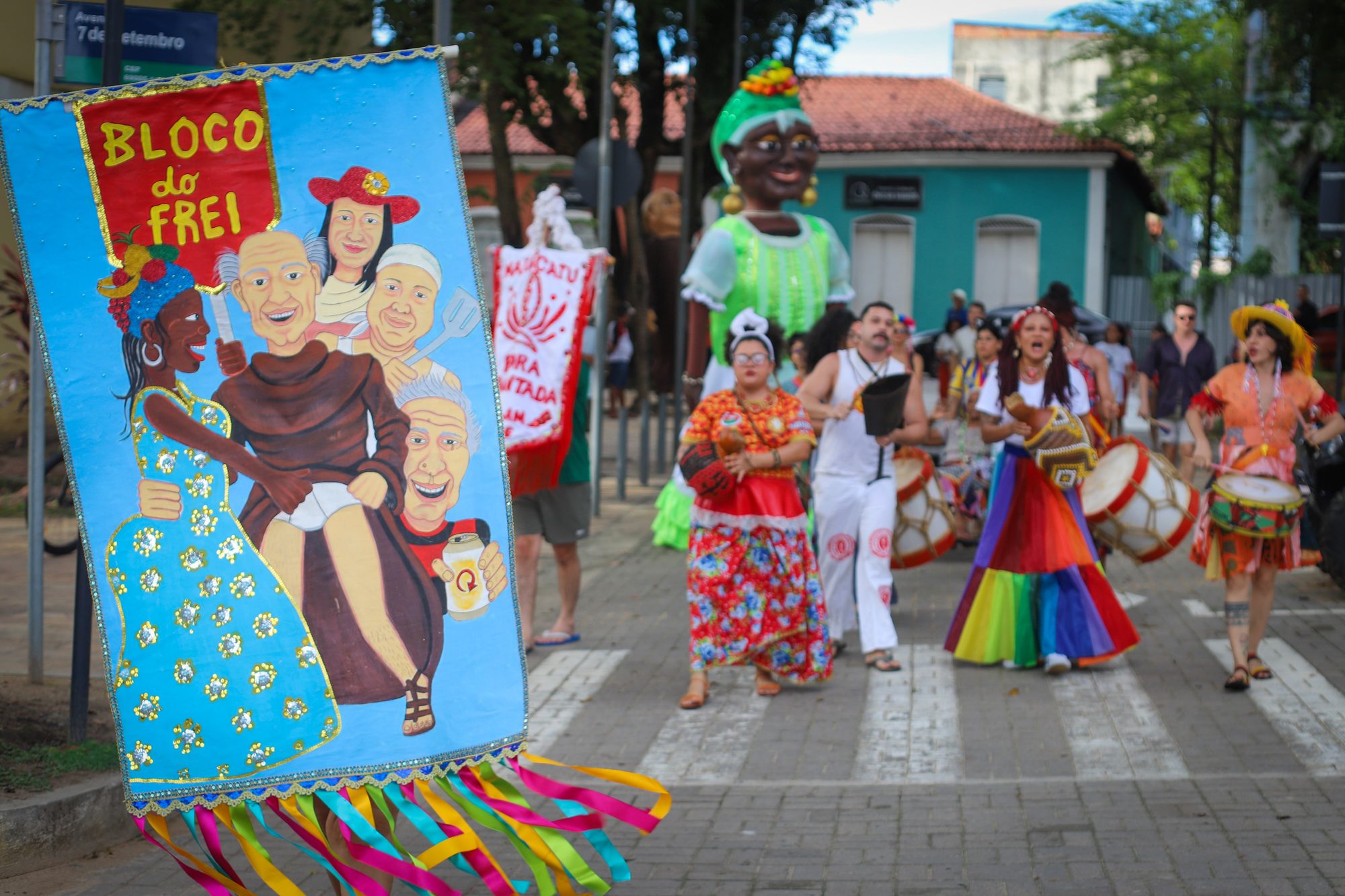 Bloco de rua no centro de Manaus durante o carnaval, com estandarte pintado à mão, boneco gigante de vestido verde, batuqueiros com tambores e a placa da avenida Sete de Setembro ao fundo.