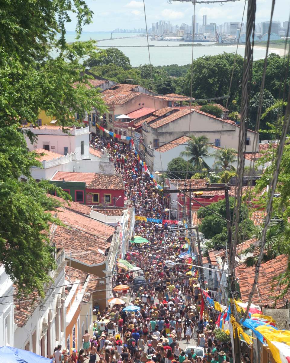 Ladeira da Misericórdia, em Olinda, lotada de foliões no Carnaval, vista do alto entre árvores e telhados coloniais, com o mar e os prédios do Recife ao fundo.