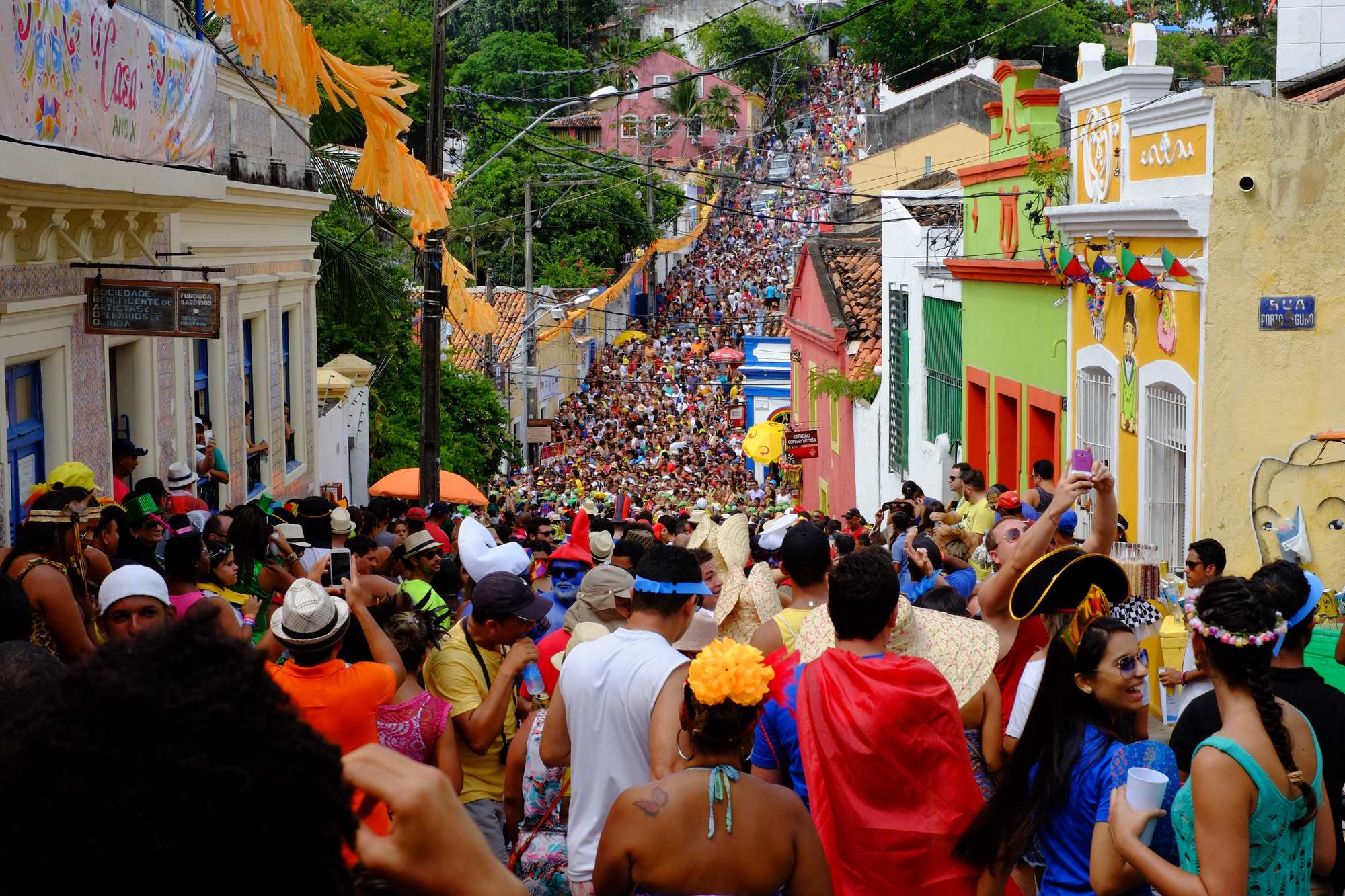 Ladeira do Sítio Histórico de Olinda tomada de foliões fantasiados, com chapéus, capas e perucas, entre casarões coloridos de amarelo, verde e rosa, bandeirolas e fitas penduradas, e a multidão subindo até o alto do morro.
