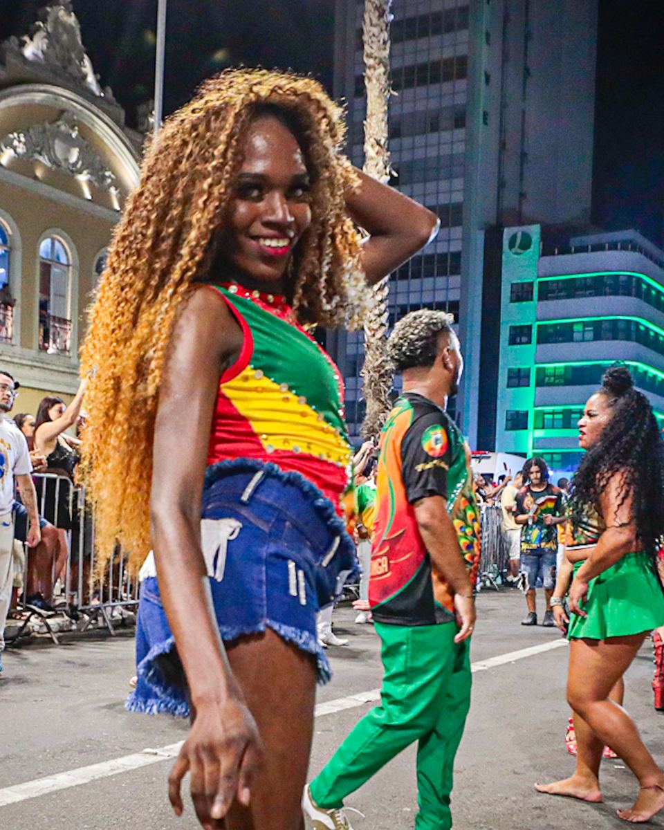 Foliã de cabelo cacheado loiro, top verde, amarelo e vermelho e short jeans, sorrindo para a câmera na Avenida Borges de Medeiros durante a Descida da Borges, no Centro Histórico de Porto Alegre, com passistas, público na grade e prédios antigos iluminados ao fundo.