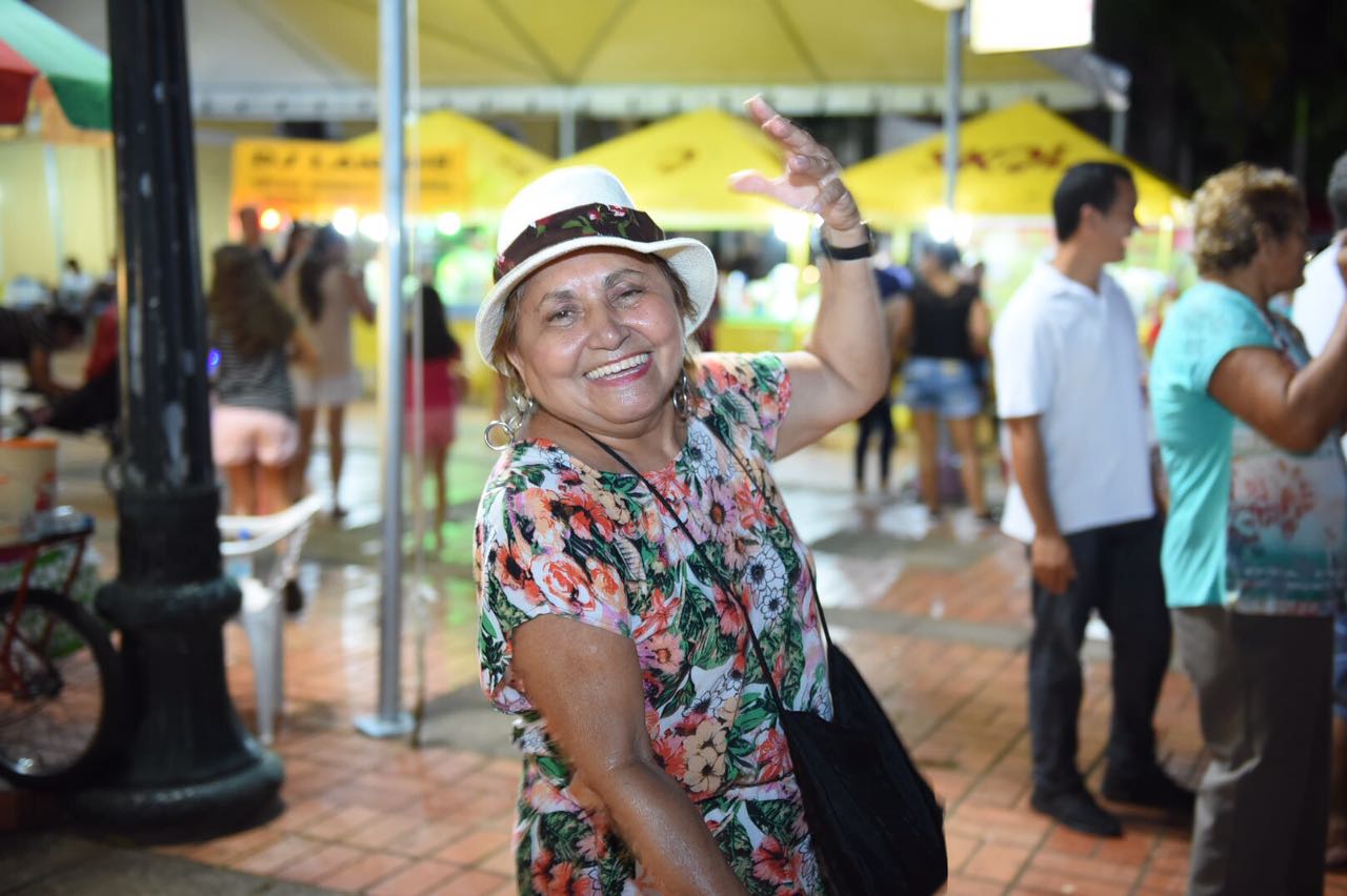 Mulher sorridente dançando com o braço erguido durante a festa de rua do Carnaval de Rio Branco, com barracas amarelas de comida e bebida iluminadas ao fundo.