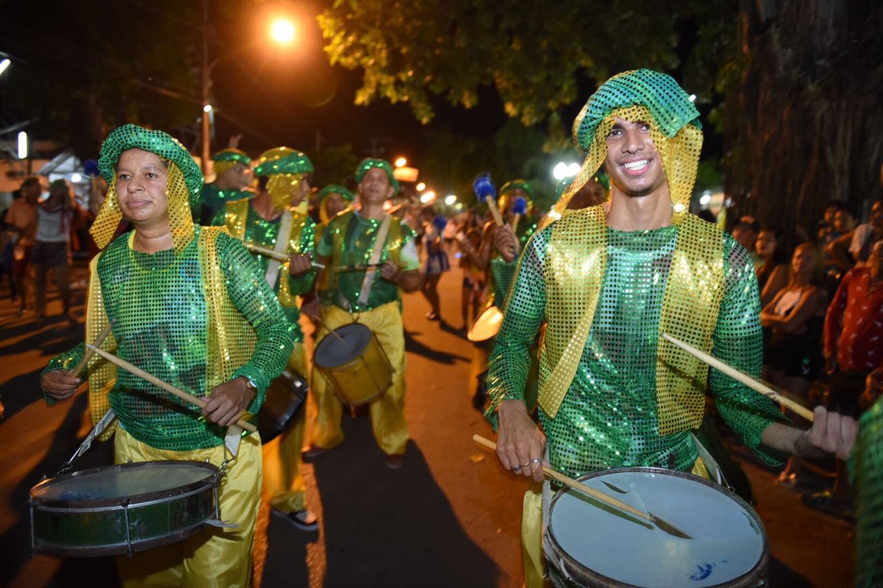 Bateria de bloco carnavalesco desfilando à noite numa rua de Rio Branco, integrantes vestidos de verde e amarelo tocando tambor, um deles sorrindo para a câmera, com o público observando dos dois lados.
