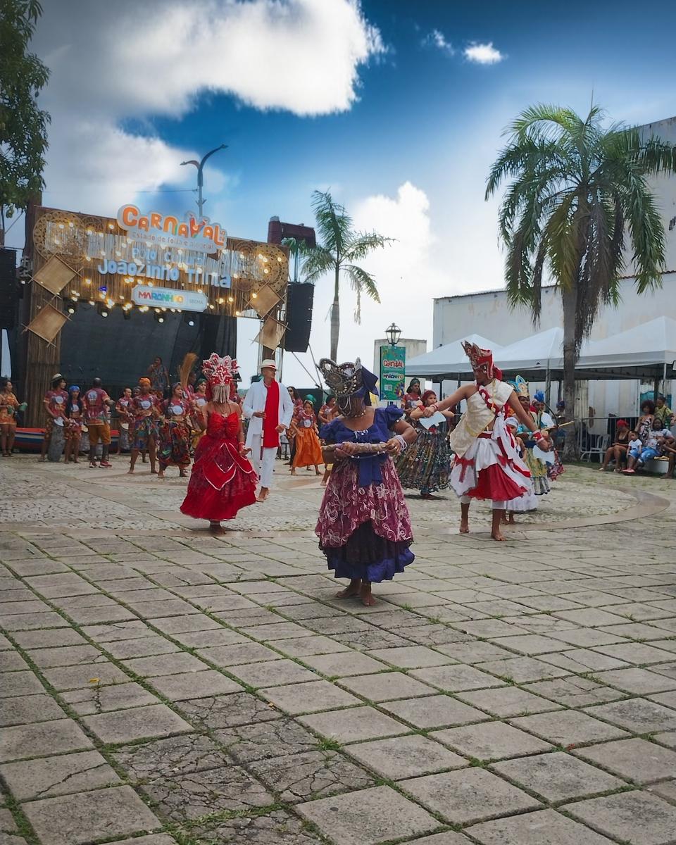 Brincantes de vestido rodado vermelho, roxo e branco e adereço de cabeça dançando numa praça de São Luís durante apresentação de bloco afro no carnaval, com palco, coqueiros e o grupo esperando a vez ao fundo.