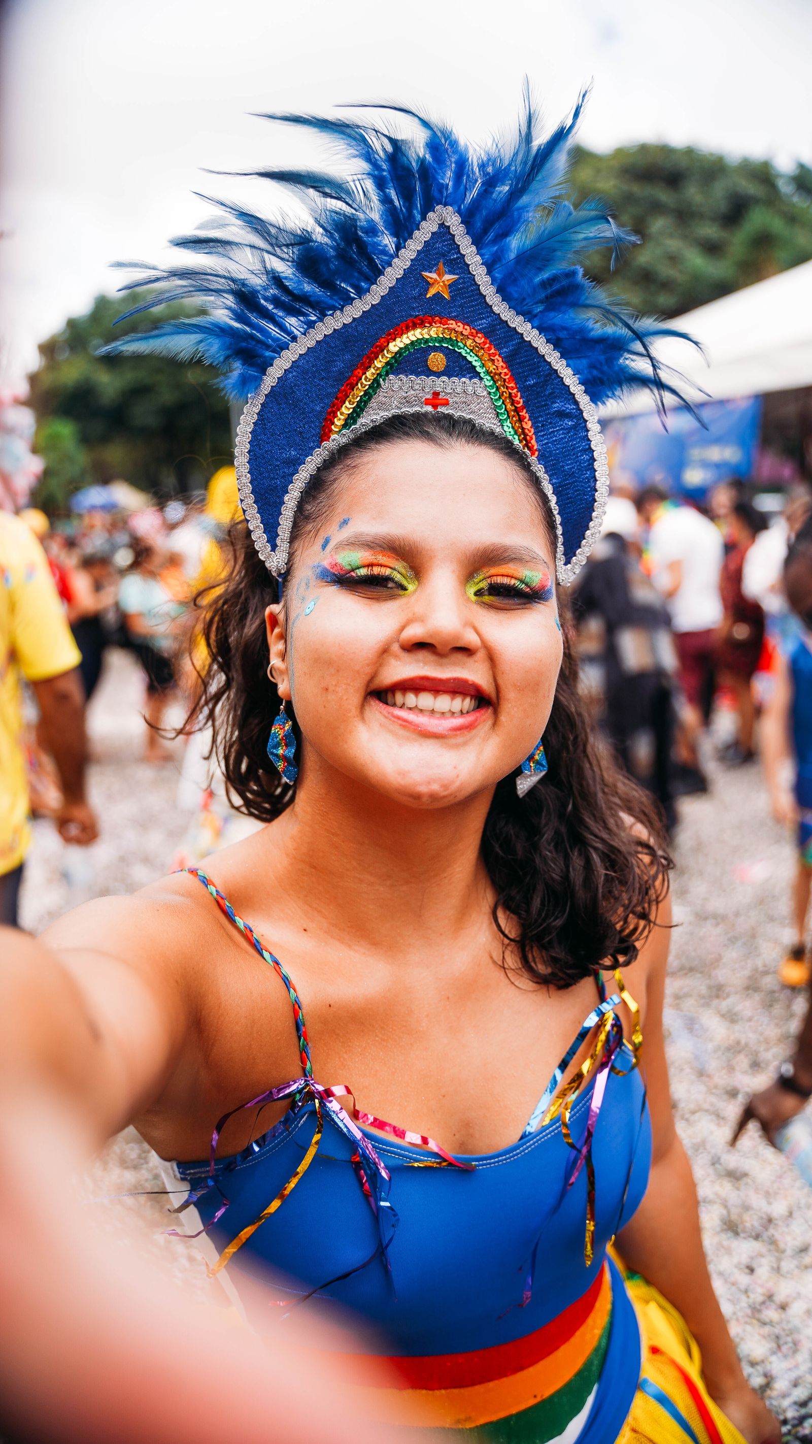 Mulher sorridente com adereço de penas azuis e maquiagem colorida tira uma selfie em meio à multidão de um carnaval de rua.