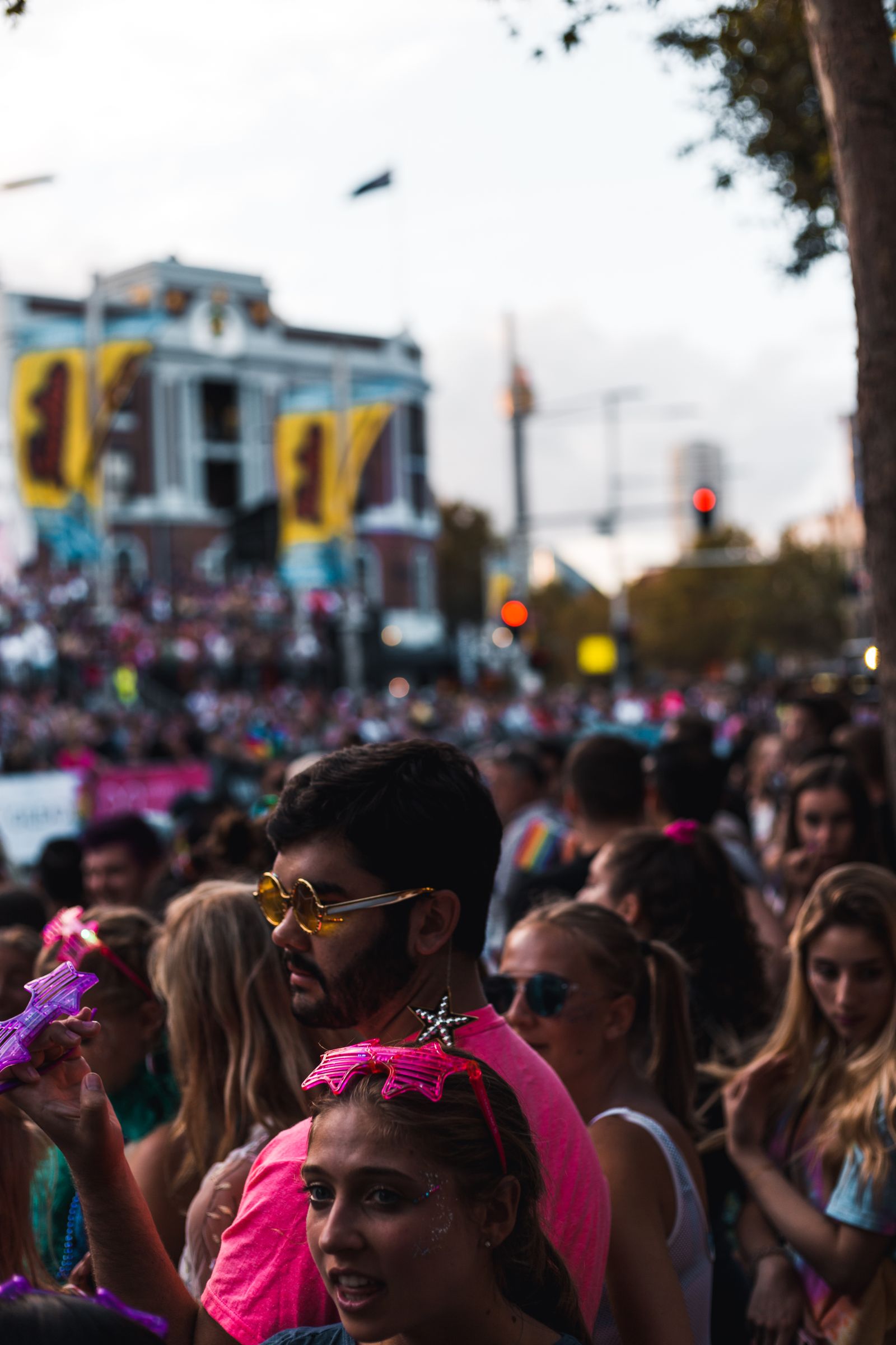 Multidão comemorando em uma rua de carnaval ao entardecer, com pessoas de braços erguidos em primeiro plano e um prédio histórico com bandeiras desfocado ao fundo.