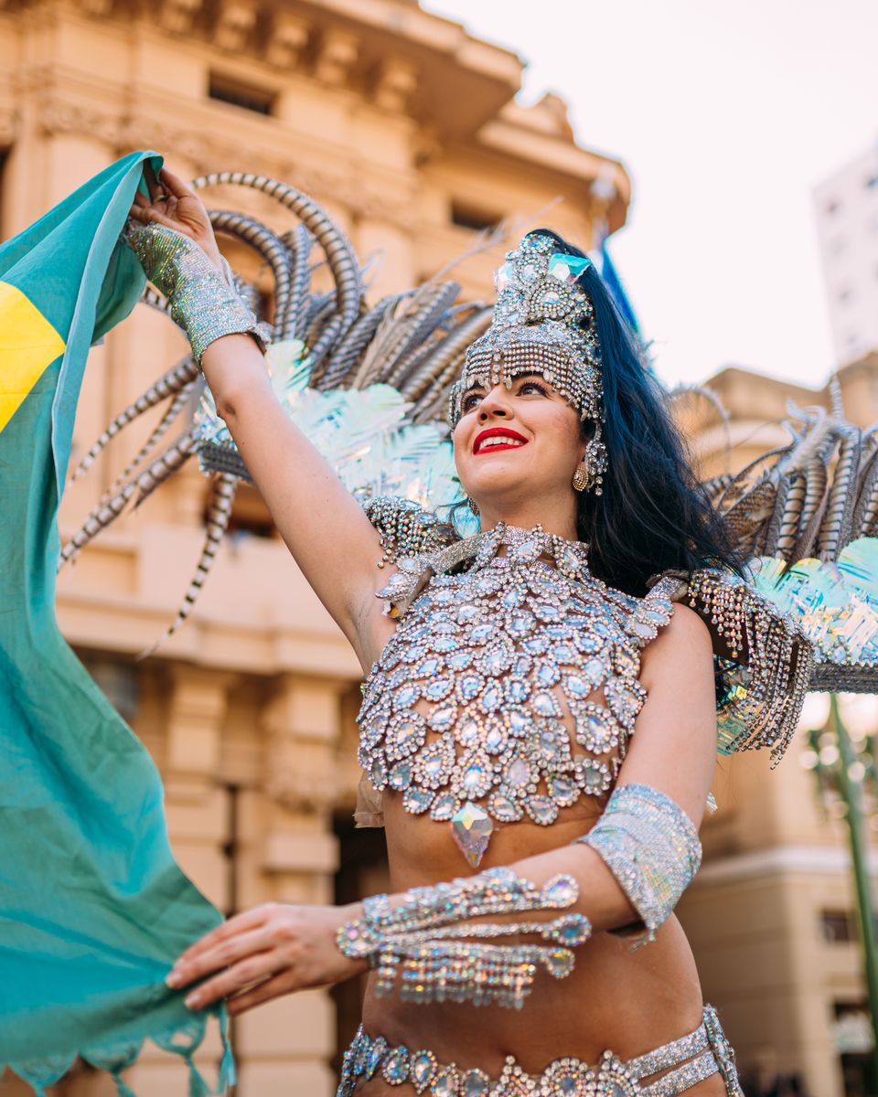 Foliã fantasiada, com adereço de plumas e bandeira do Brasil, sorrindo num desfile de Carnaval de rua.