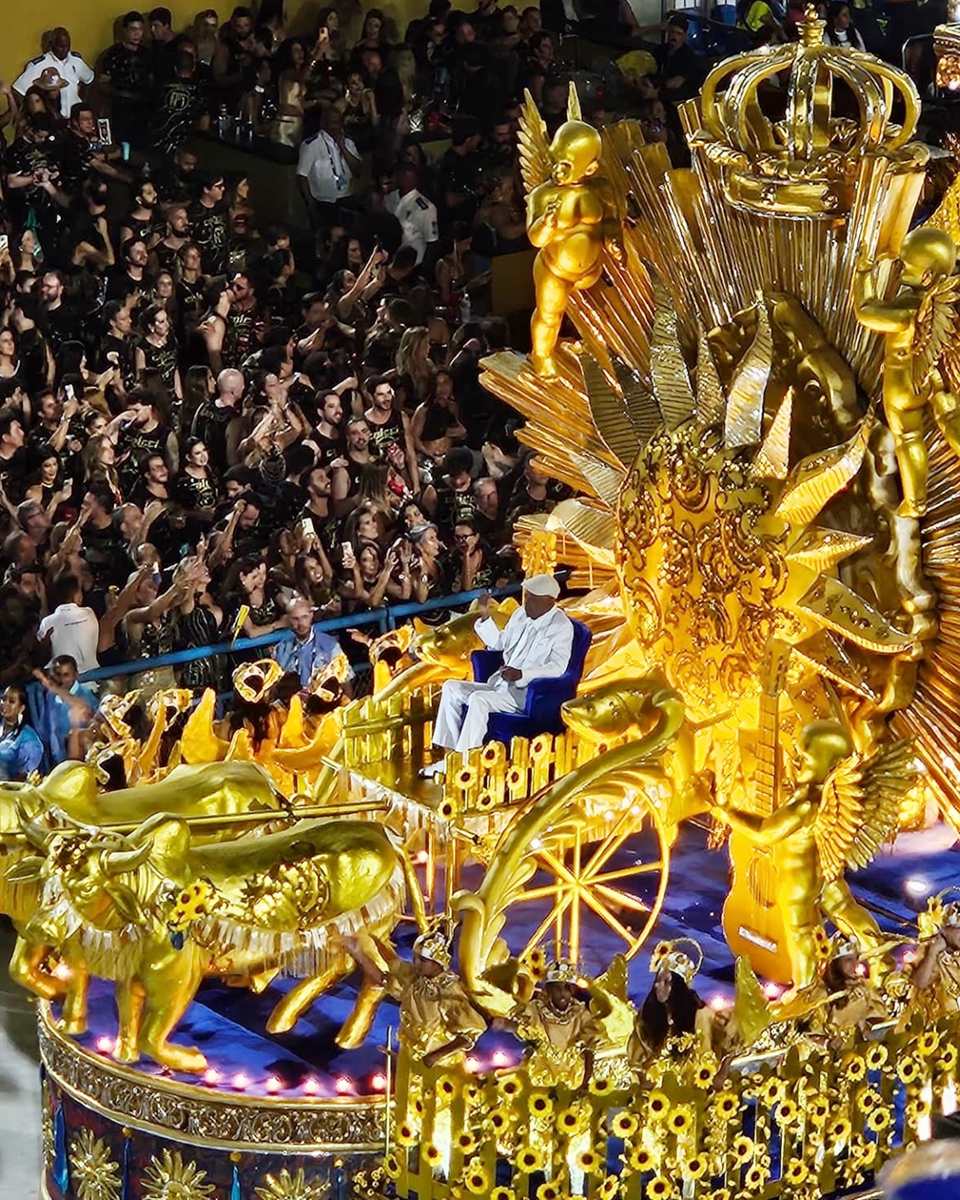 Carro alegórico dourado, com anjos, coroa e sol esculpidos, passando diante da arquibancada lotada do Sambódromo, com muita gente filmando com o celular, no Desfile das Campeãs.
