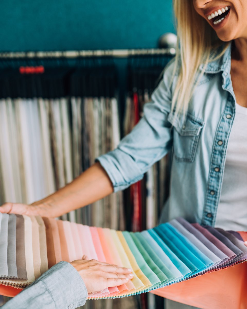Mulher loira de camisa jeans sorrindo e abrindo um mostruário de tecidos coloridos para cortina, numa loja de tecidos com rolos pendurados ao fundo.