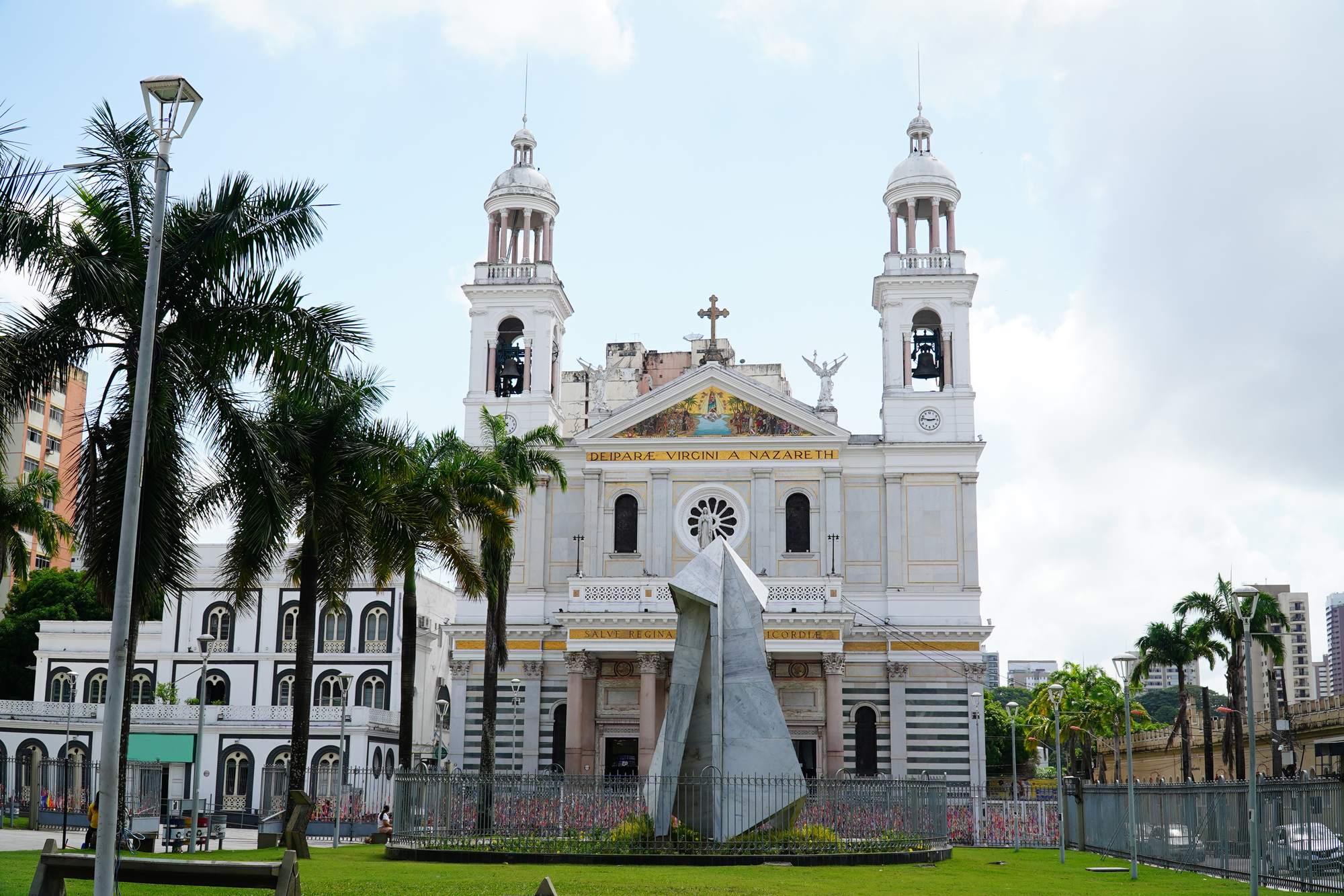 Fachada branca da Basílica Santuário de Nossa Senhora de Nazaré, em Belém, com as duas torres, palmeiras e o monumento da Praça Santuário em primeiro plano.