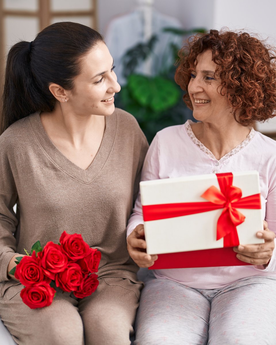Mãe de cabelo cacheado e filha adulta sentadas na cama, sorrindo uma para a outra, com uma caixa de presente branca de laço vermelho e um buquê de rosas vermelhas.