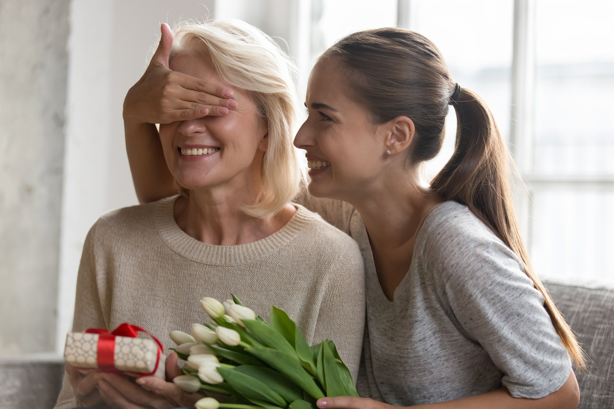 Filha adulta tapa os olhos da mãe, sorridente no sofá da sala, com uma caixa de presente e um buquê de tulipas brancas no colo.