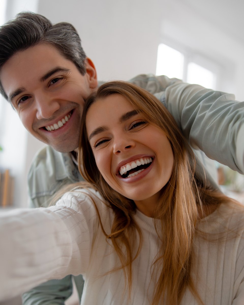 Casal jovem rindo e fazendo selfie com o celular na sala de casa, com um vaso de rosas vermelhas e um balão de coração ao fundo.