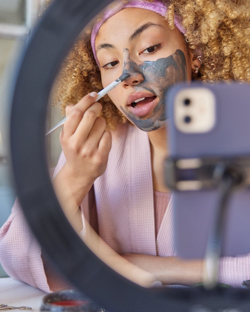 Jovem de cabelo cacheado e roupão rosa aplicando máscara de argila cinza no rosto com um pincel, vista através de um ring light, enquanto grava com o celular preso num suporte.