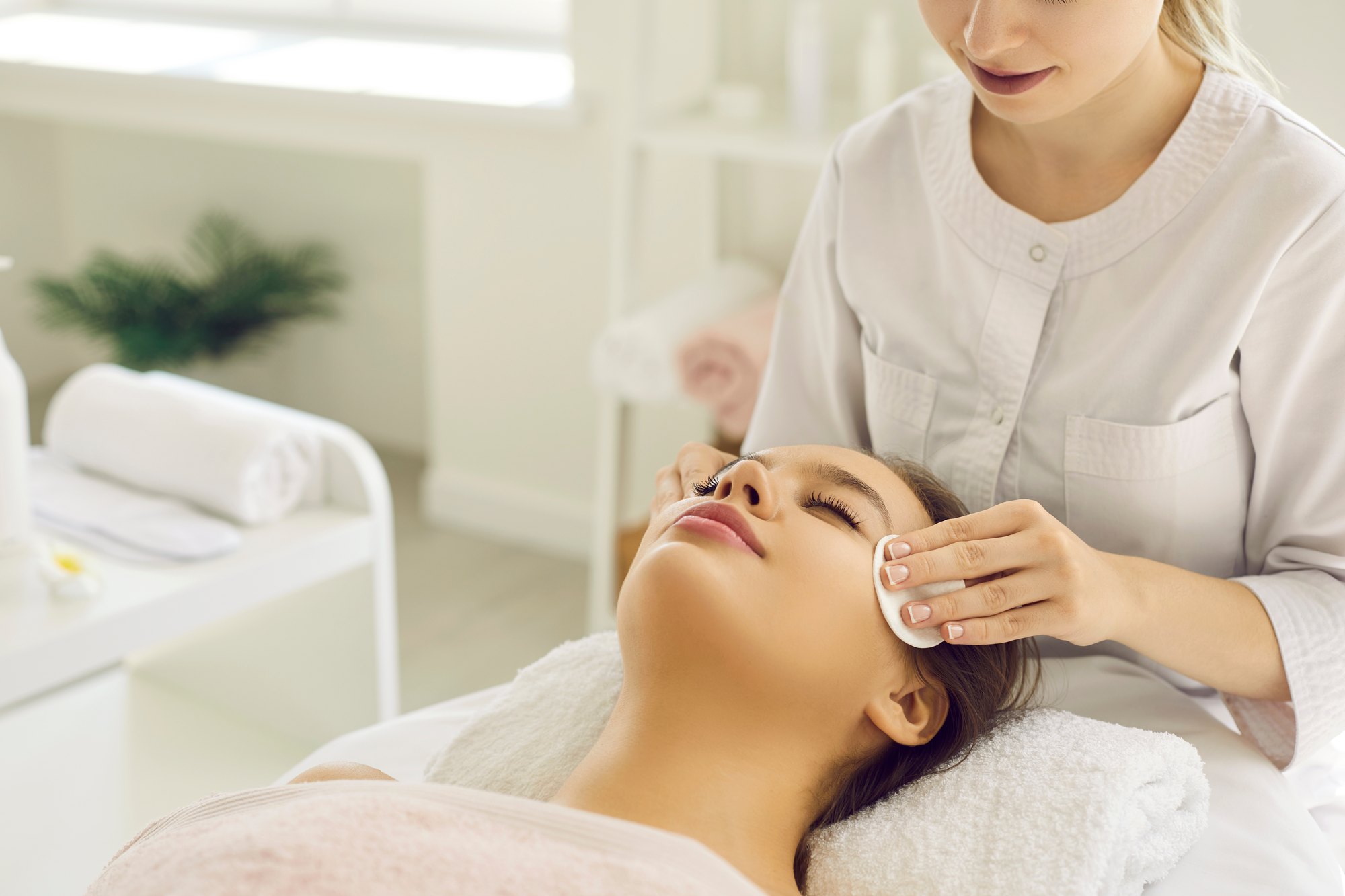 Esteticista de jaleco branco limpando com disco de algodão o rosto de uma cliente deitada numa maca, numa cabine de estética clara com toalhas enroladas ao fundo.