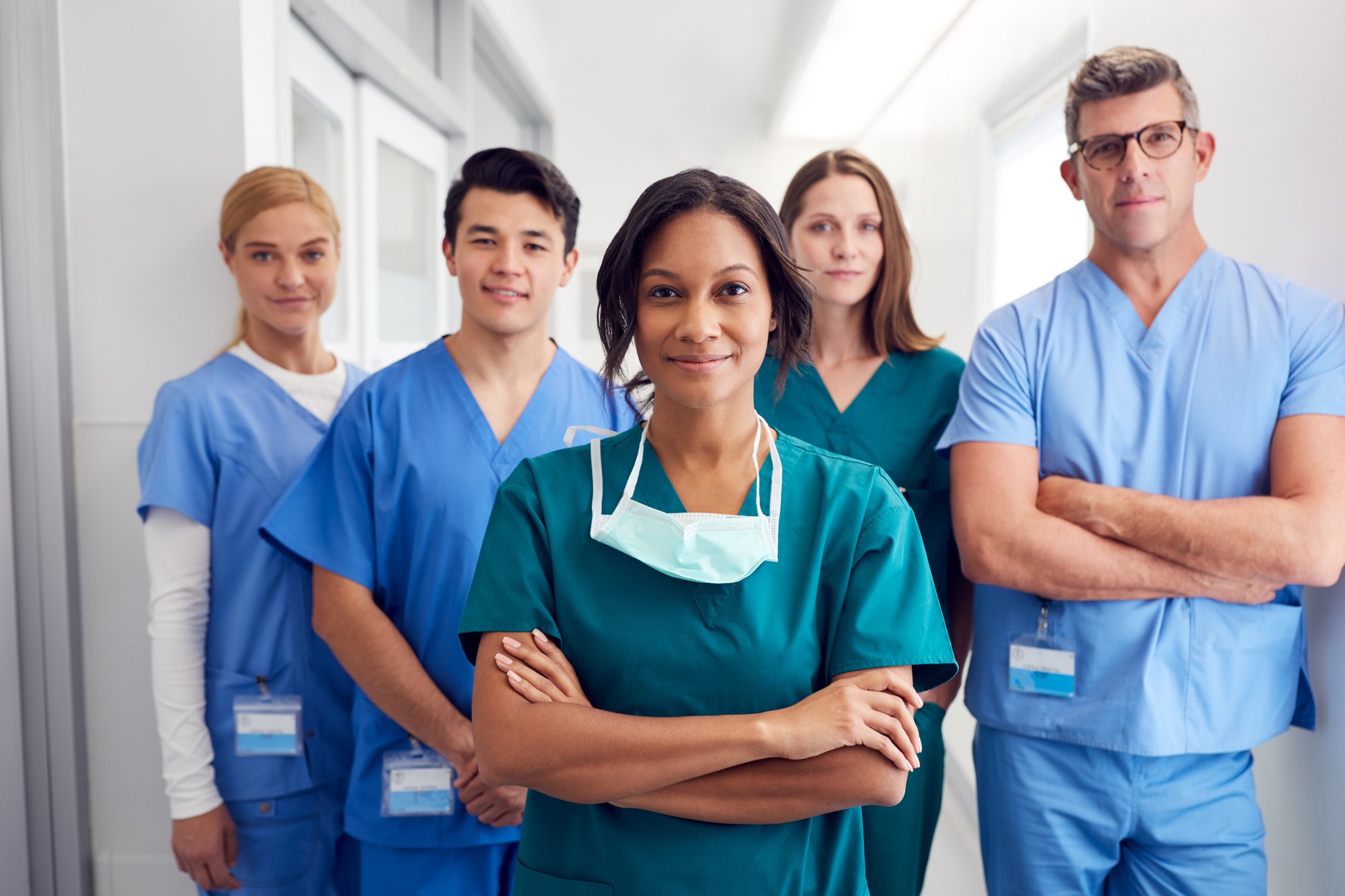 Equipe de cinco profissionais de saúde de pijama cirúrgico azul e verde, com crachás, parada num corredor claro de hospital; à frente, uma mulher de braços cruzados com a máscara abaixada no pescoço.