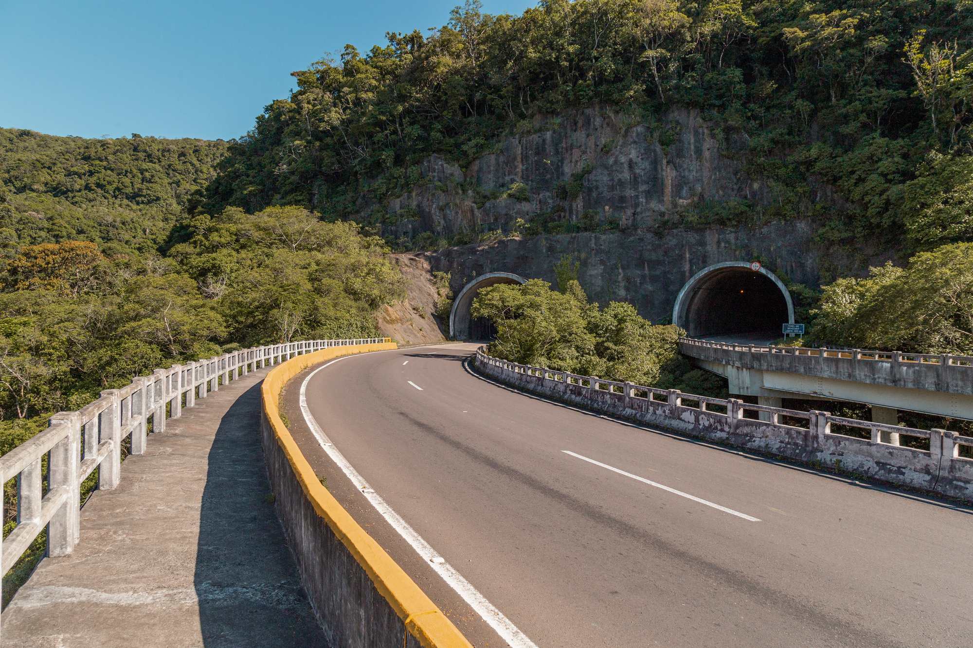 Rodovia Rota do Sol, na serra do Rio Grande do Sul, com curva sobre um viaduto de mureta amarela, dois túneis abertos na rocha e mata atlântica cobrindo a montanha num dia de céu azul.